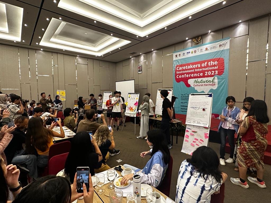 A group of people attending a conference titled 'Caretakers of the Environment International Conference 2023' in a large, well-lit room. Some individuals are giving presentations at the front with posters and speaker, while others are seated at tables, taking photos and listening.