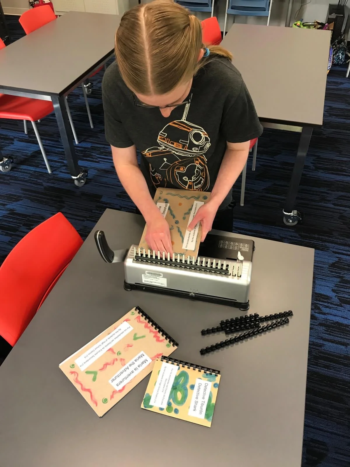 A person is using a paper punch machine at a table, punching holes into a paper with a map design. There are colorful educational booklets on the table, with one titled 'Detective Tutori Detective Stark.' The room has a blue striped carpet and red ch