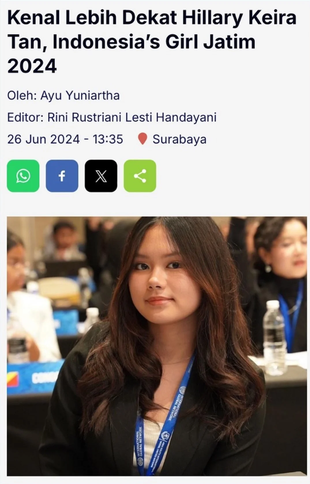 A young woman with medium-length brown hair, wearing a black blazer and a conference badge around her neck, sitting at a conference table with blurred people in the background.