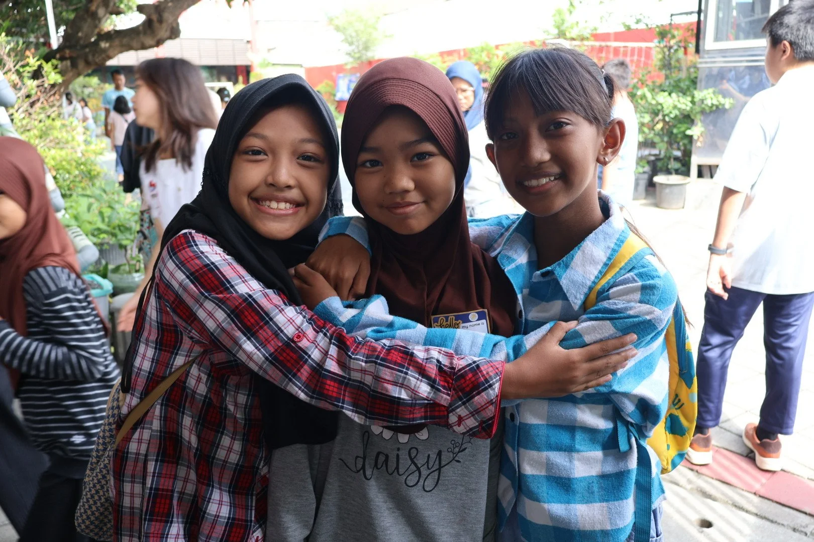 Three young girls in colorful shirts and headscarves hugging and smiling at the camera in an outdoor setting with other people and greenery in the background.