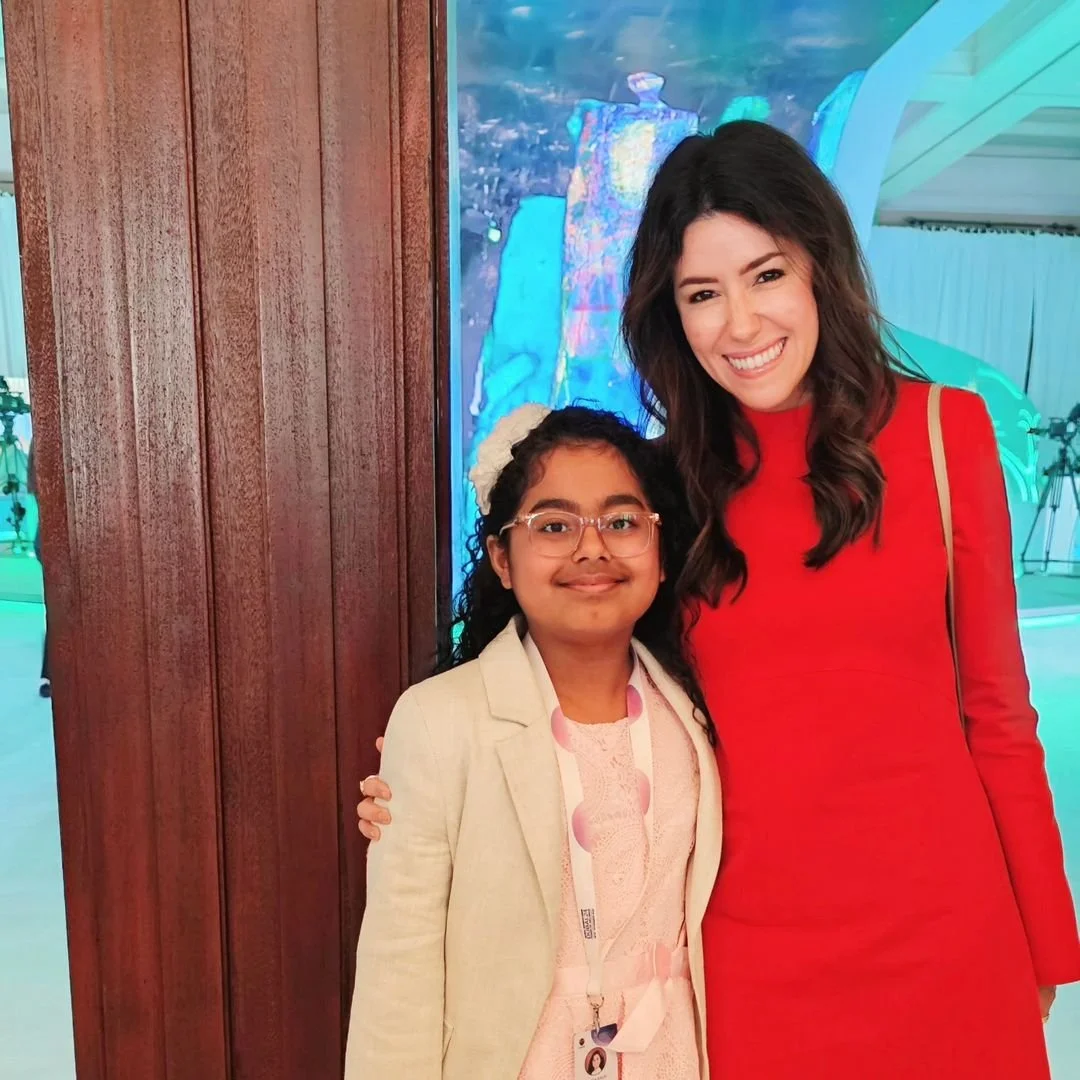A woman in a red dress standing next to a young girl in a beige blazer and glasses, both smiling for the photo indoors with colorful lights in the background.