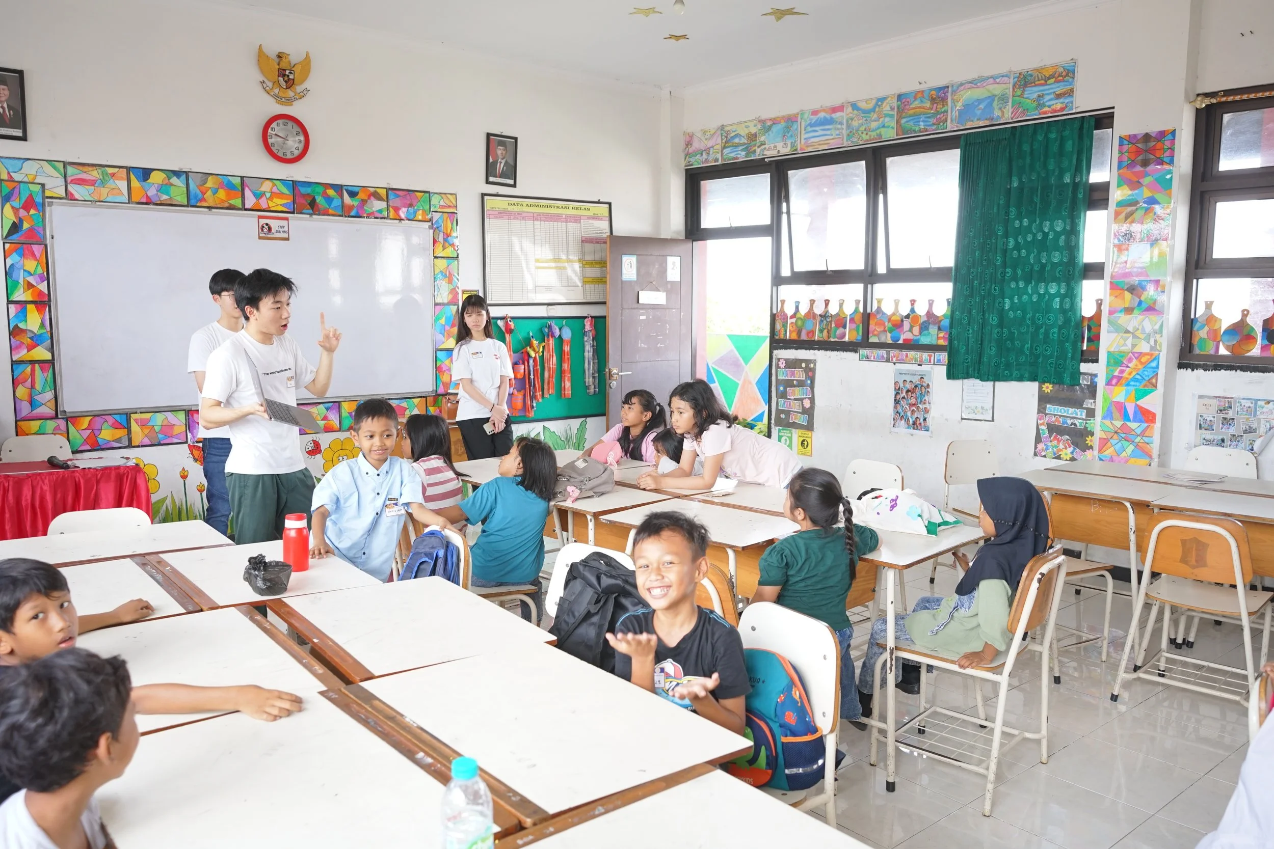 A classroom filled with young students, some seated at desks and others standing, with teachers or students speaking at the front. The classroom is decorated with colorful artwork and educational posters, with large windows letting in natural light.