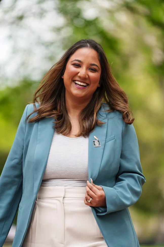 A woman with long brown hair smiling outdoors, wearing a light blue blazer and white top, with blurred green trees in the background.
