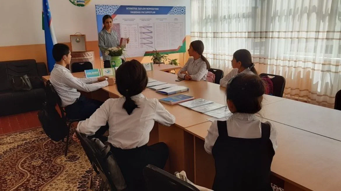 A woman presenting at a conference table with five children or young students sitting around it, in a room with a large chart on the wall and large windows with curtains.
