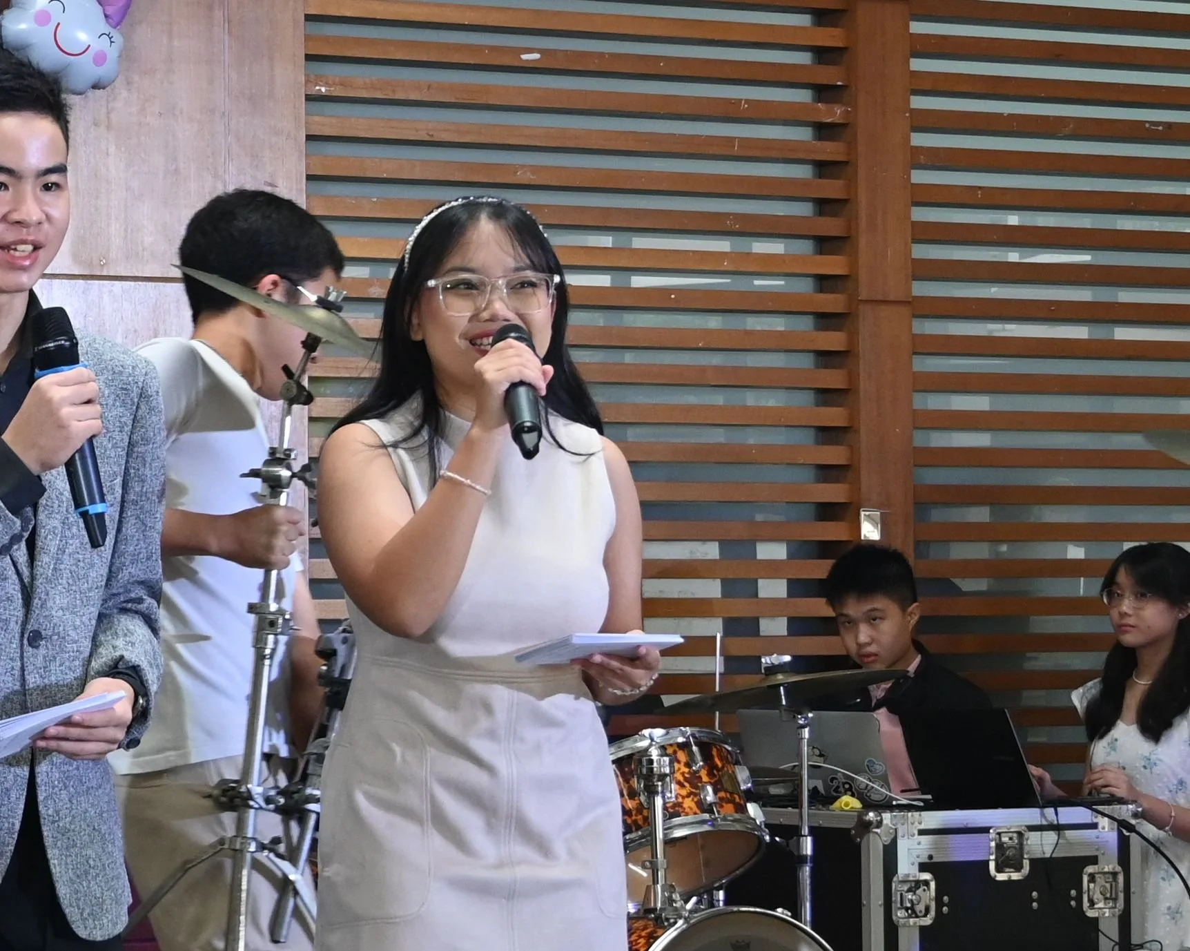 A group of people at an indoor event. One woman holding a microphone and a notepad, smiling and speaking. Two men and one woman, some holding microphones or notes. There is a drum set and musical equipment in the background. Wooden slatted wall behind them.