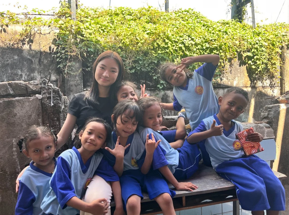 A group of children and a young woman posing together outdoors. The children are in school uniforms, some making fun gestures, and the woman is smiling. They are sitting and standing on a bench with a stone wall and greenery in the background.