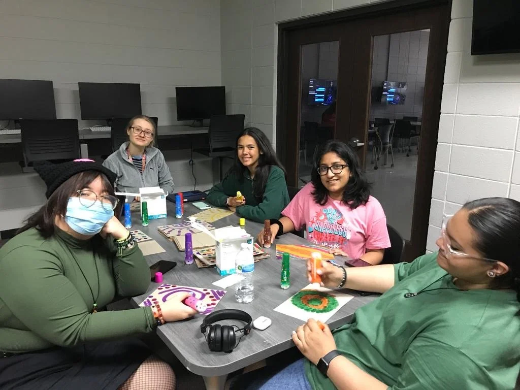 Group of six women sitting around a table in a computer lab with computers on the wall behind them, engaged in arts and crafts activities.