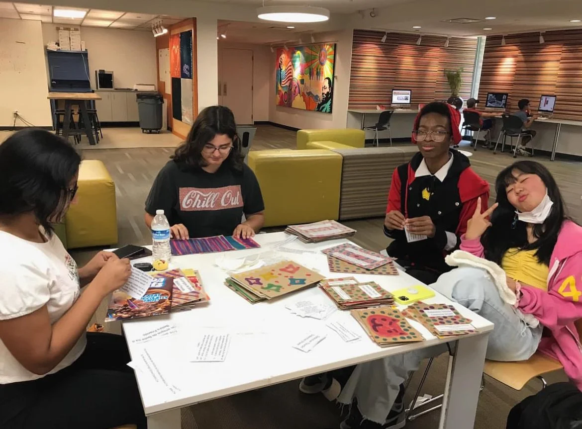 Four young women sitting at a table with various craft supplies, in a room with colorful artwork on the wall and computers in the background.