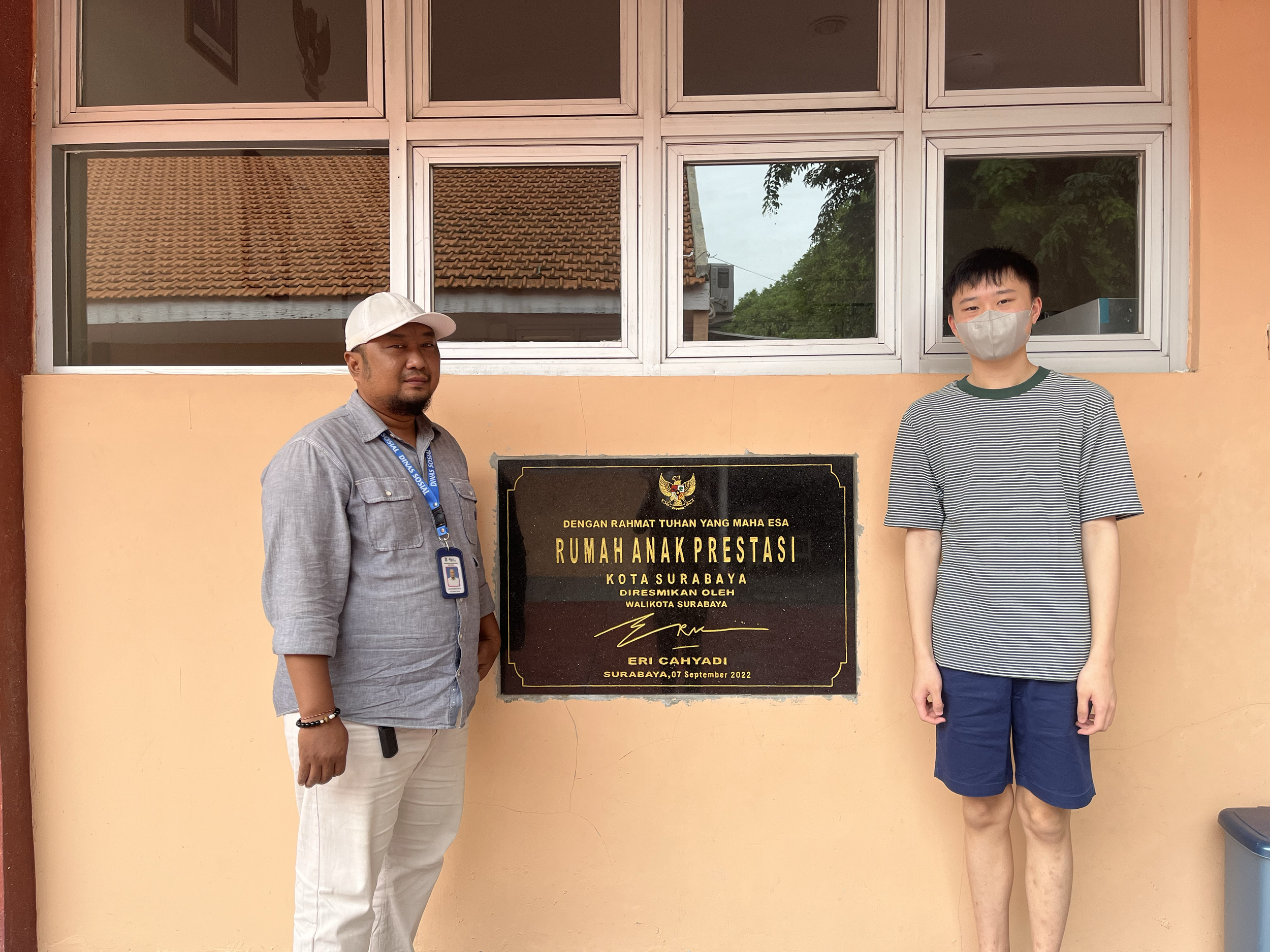 Two men standing next to a black commemorative plaque on a peach-colored wall. The plaque is inscribed with Indonesian text and a signature, indicating the dedication of a children's achievement house in Surabaya.