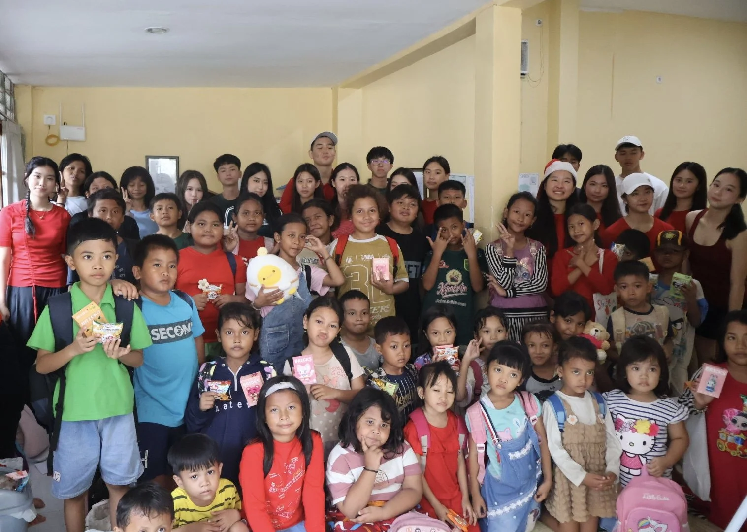 Group of children and adults gathered indoors for a group photo, some children holding toys or snacks, with many smiling and making peace signs, in a festive or celebratory setting.