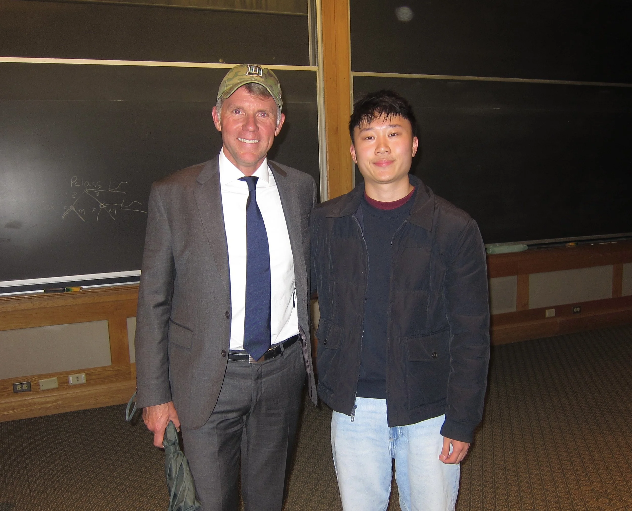 Two men standing side by side in a classroom with chalkboards in the background. One man is dressed in a suit with a tie and the other is wearing casual clothes with a jacket.