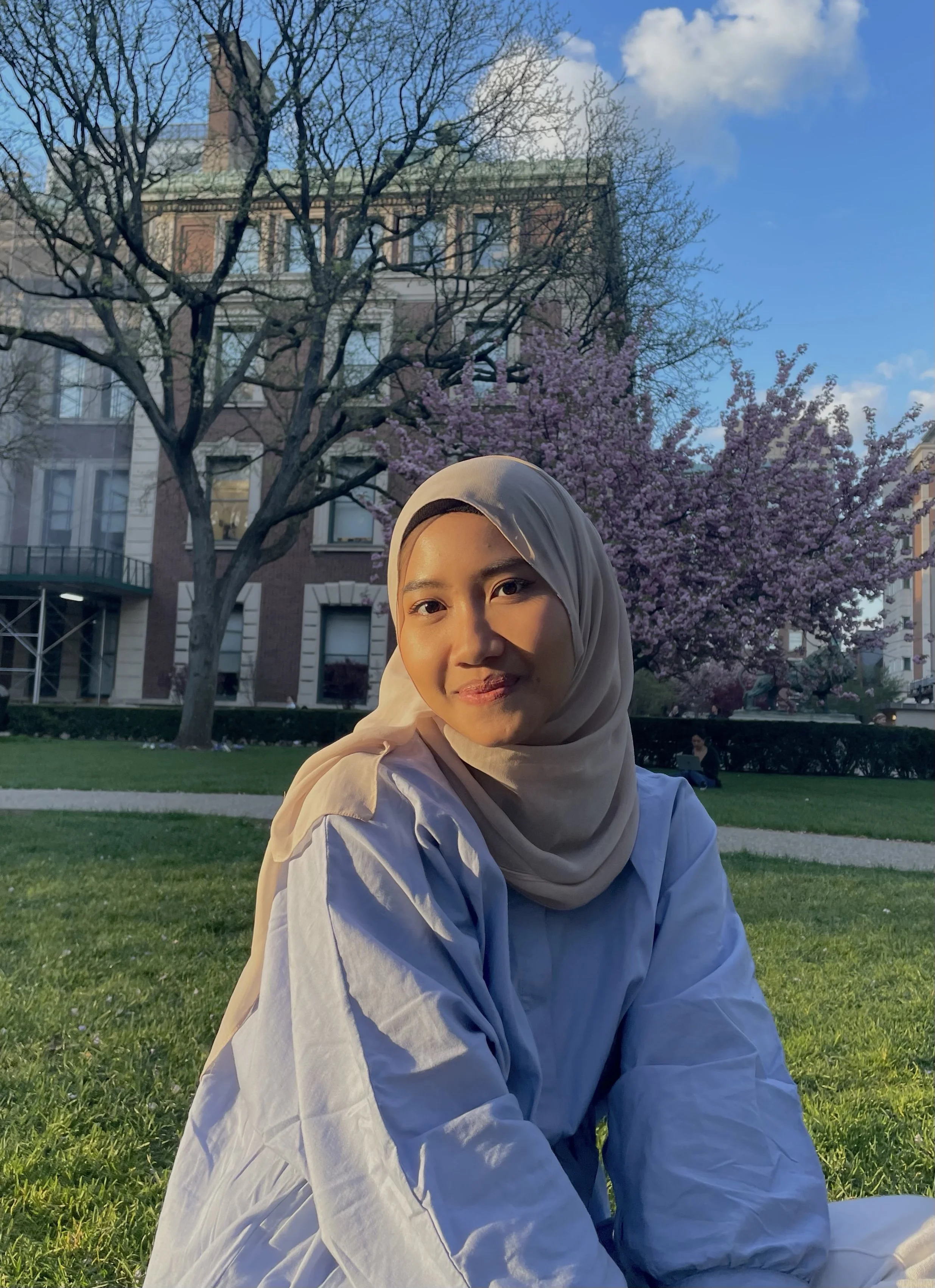 A woman wearing a beige hijab and white shirt sitting on grass in a park with blossoming pink cherry trees and a brick building in the background during late afternoon or early evening.