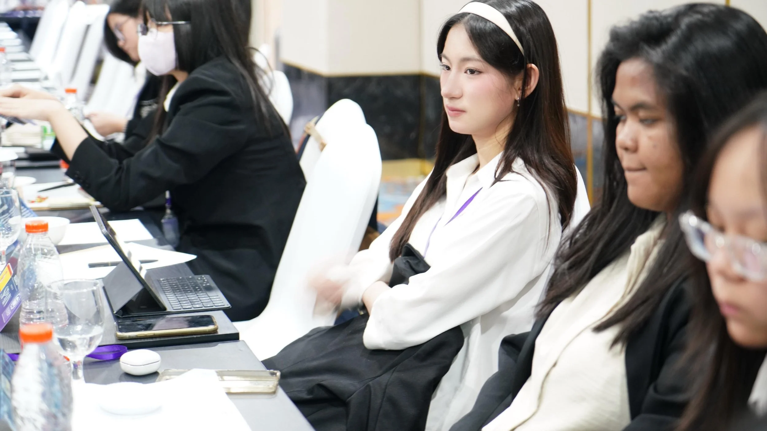 Young women sitting at a conference table, some taking notes or using laptops, with one woman in the center listening attentively.