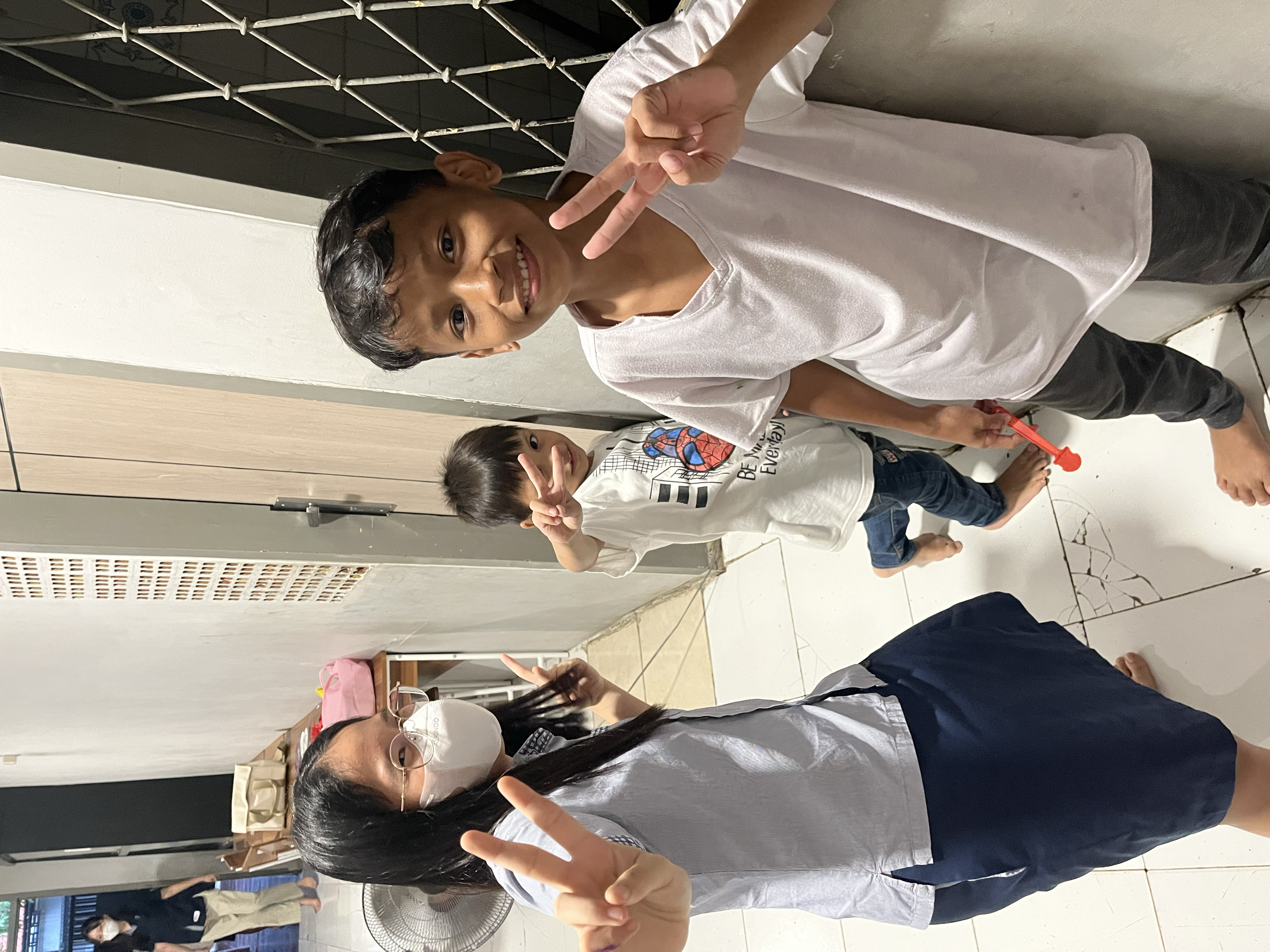 Two children and an adult indoors making peace signs with their fingers. The children are smiling, and the adult is wearing glasses and a face mask. The floor is tiled, and there are some bags and household items in the background.