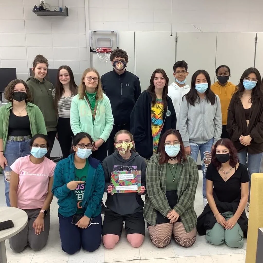 A group of fifteen diverse students wearing masks, indoors in a classroom or school setting, with some holding a colorful certificate or award.