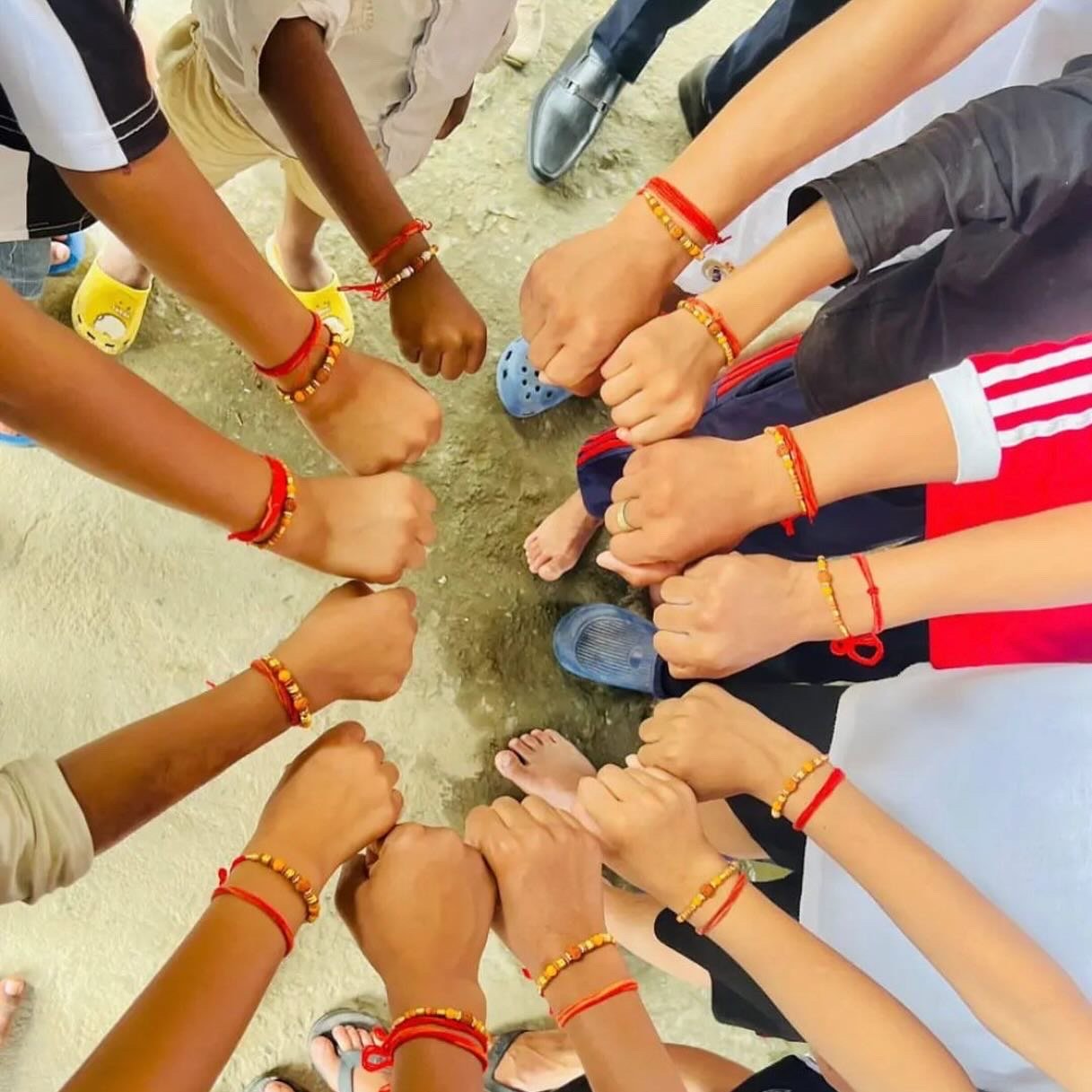 A group of children standing in a circle with their fists extended inward, wearing colorful bracelets, and standing on a dirt ground.