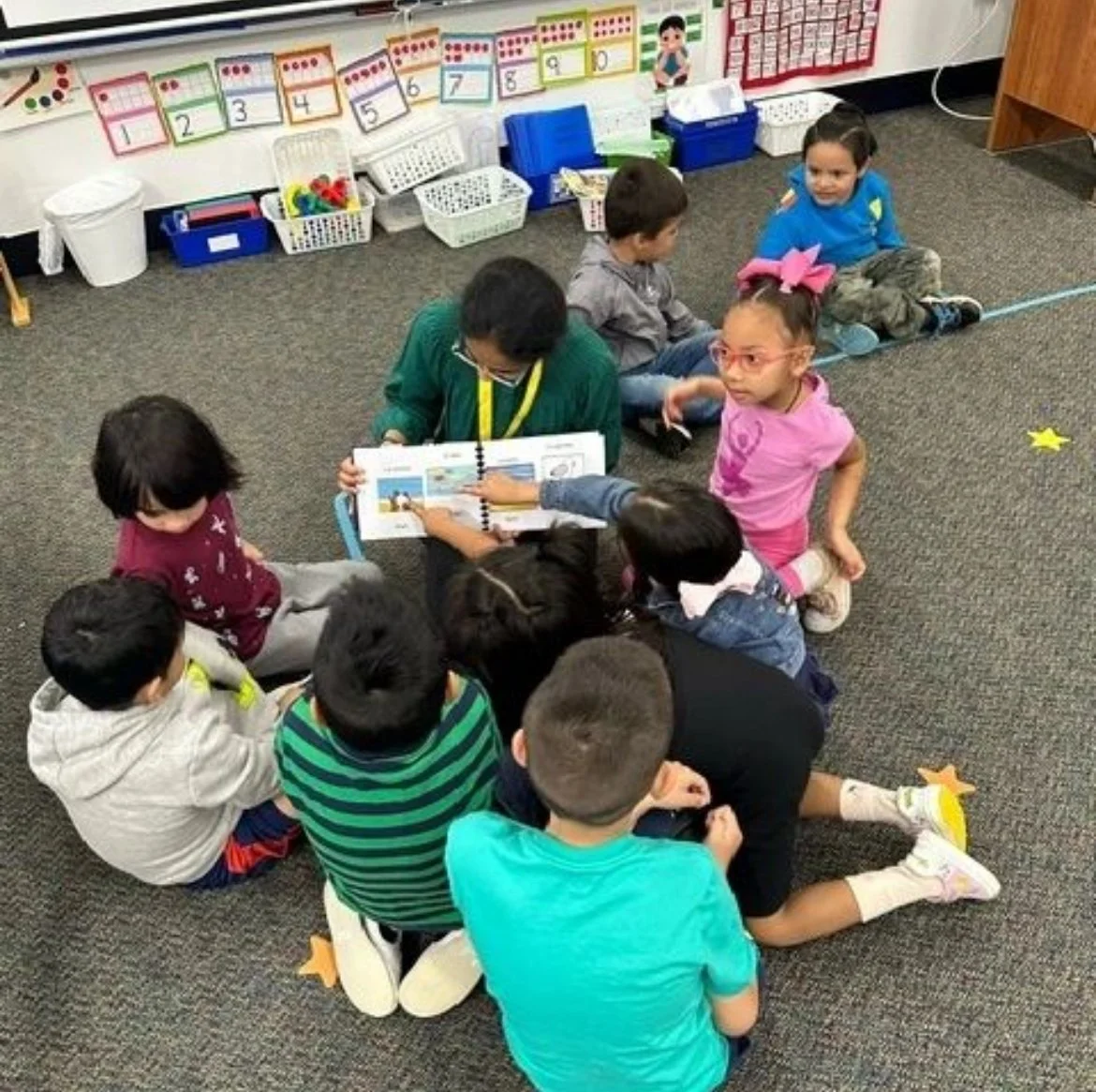 Group of young children sitting on the classroom carpet, looking at a teacher's book, with classroom bins and educational posters in the background.