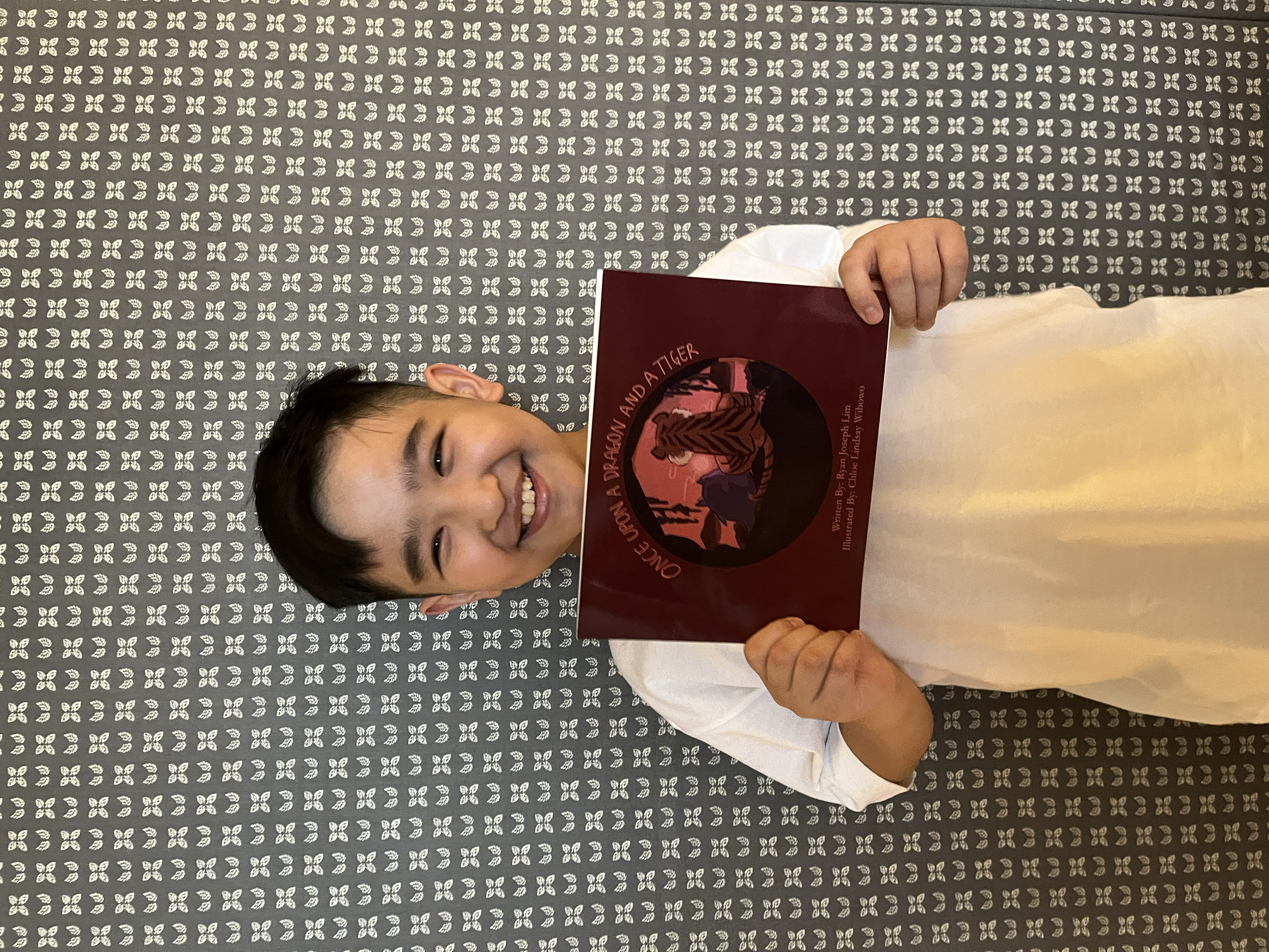 A young boy is lying on a patterned carpet, holding a book titled 'Once Upon a Dragon and a Tiger,' and smiling at the camera.