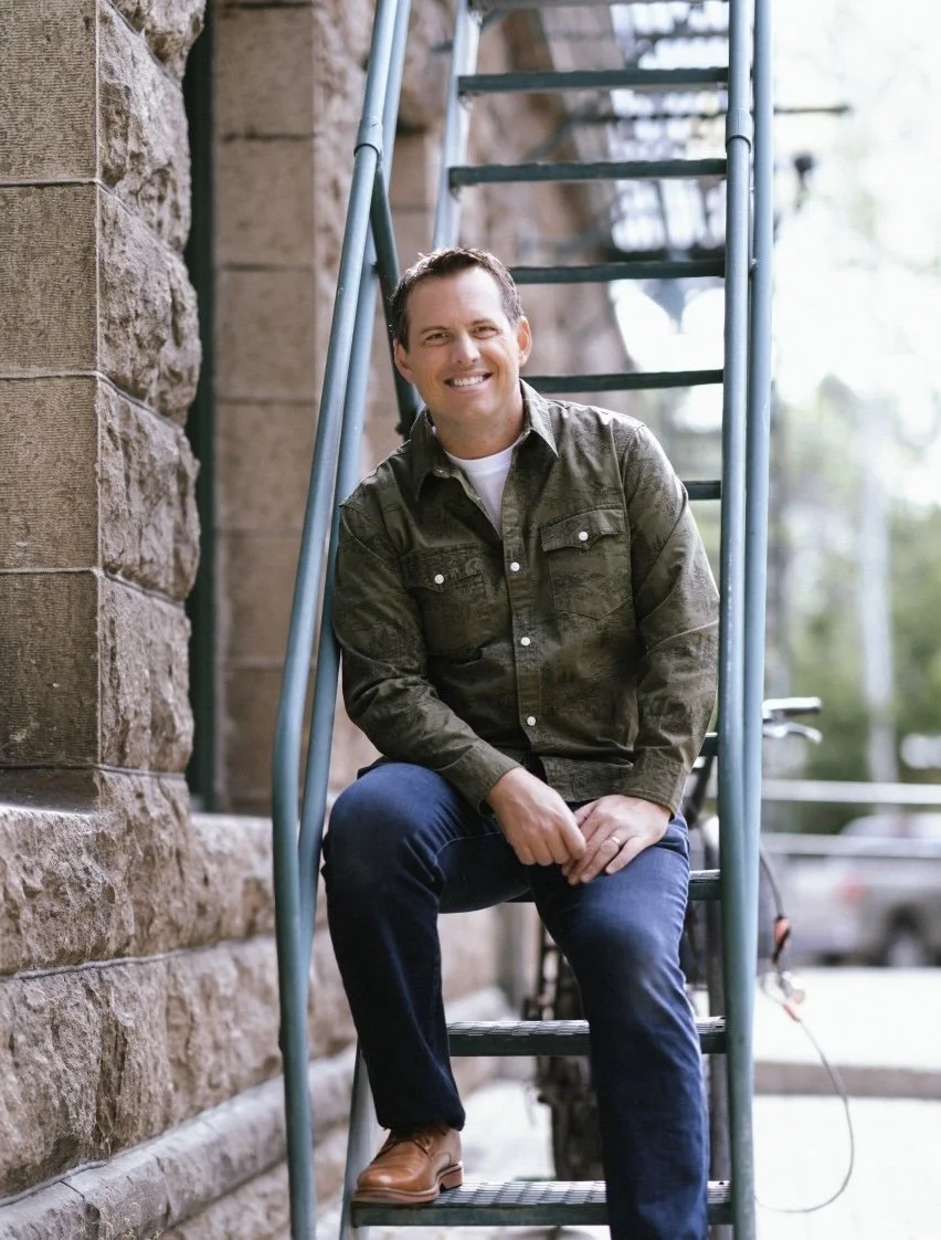 A man sitting on a ladder outside near a stone wall, smiling at the camera. He is wearing a green jacket, white shirt, dark jeans, and brown shoes.
