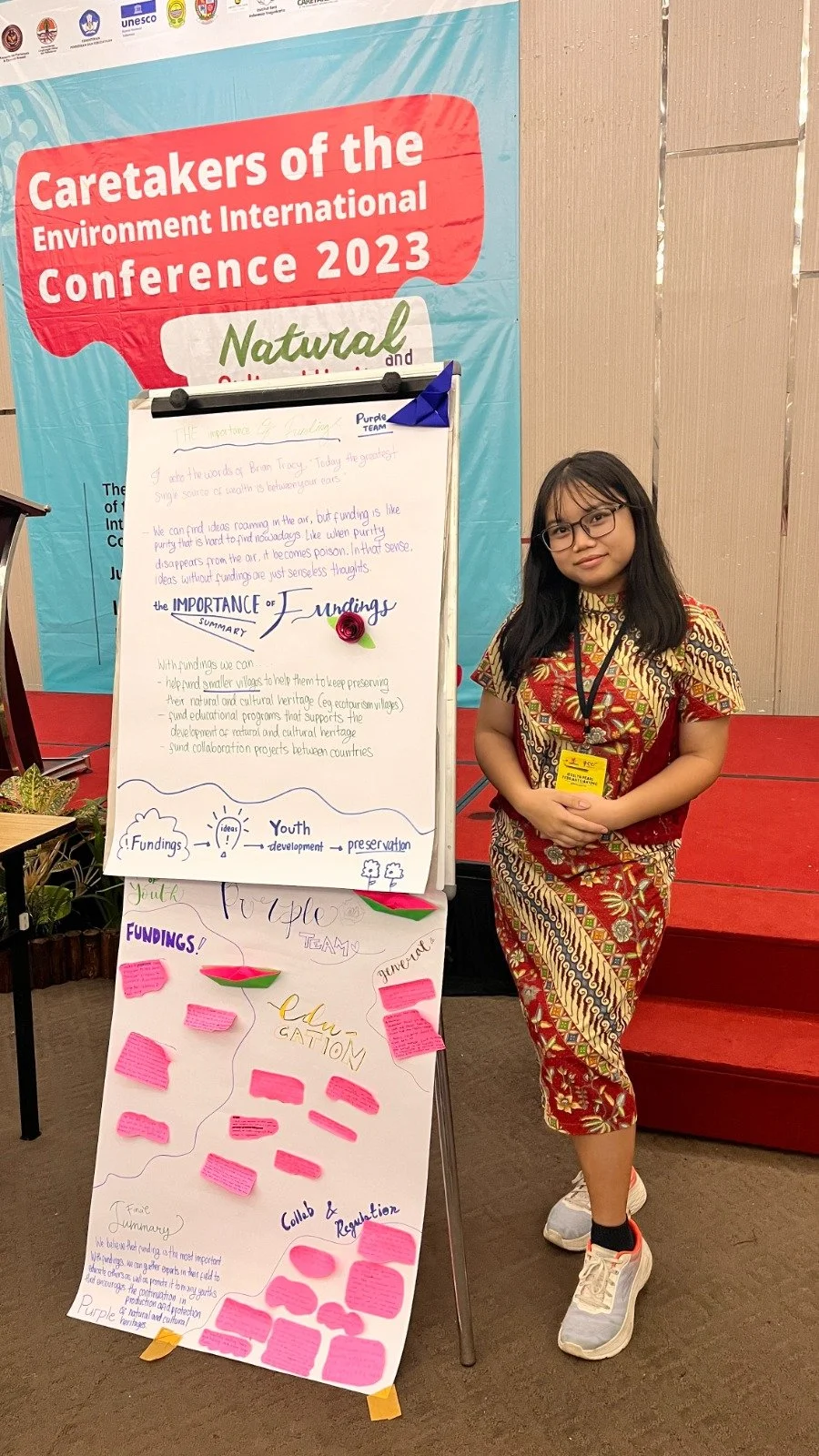 A young woman in glasses wearing a traditional batik dress, standing next to a presentation board at the 2023 Environment International Conference. The board features notes and diagrams about funding, youth development, and cultural preservation.
