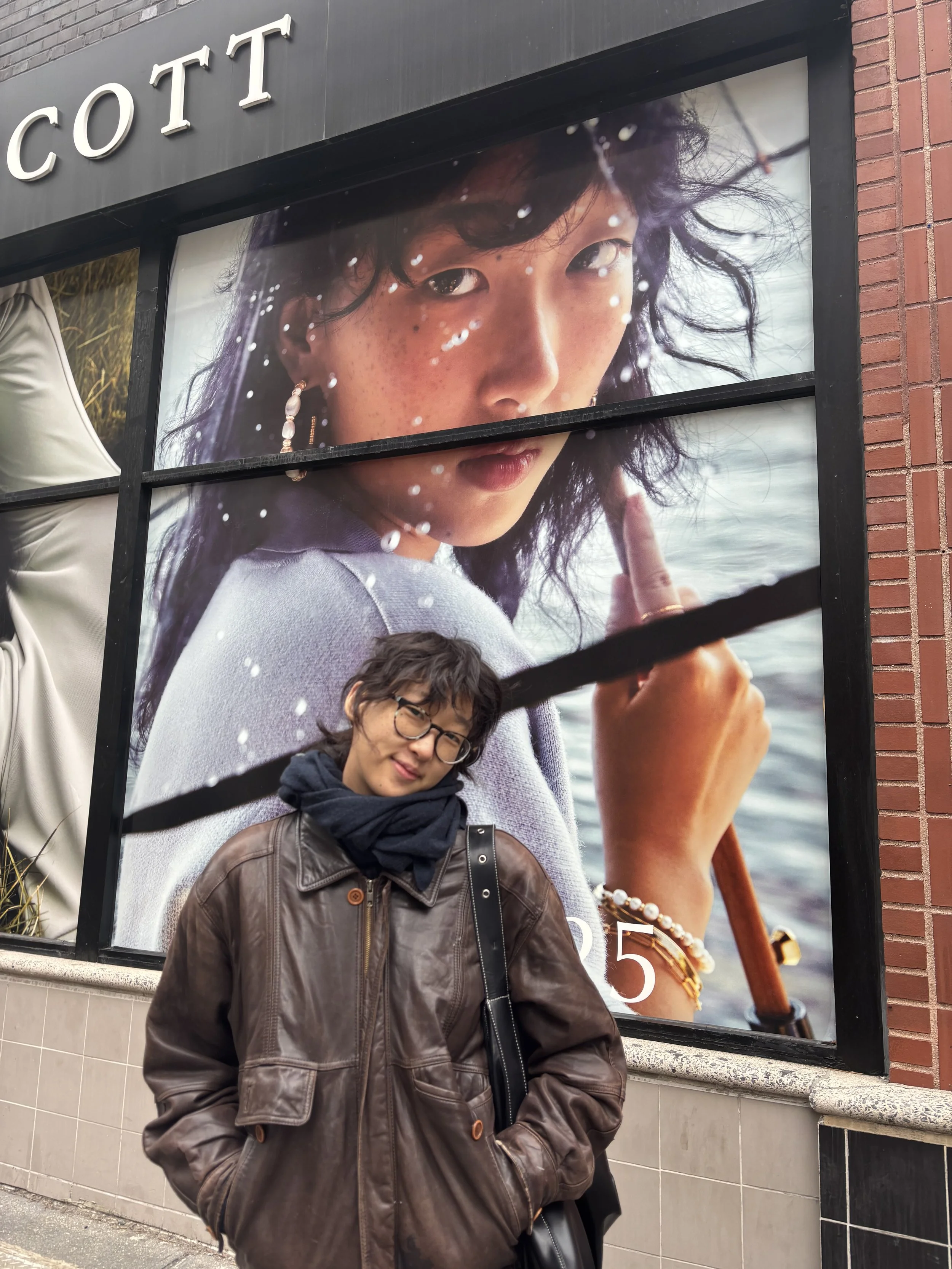 A woman with glasses and a leather jacket standing in front of a large outdoor advertisement featuring a woman with dark hair, freckles, and jewelry, holding a paddle near water.