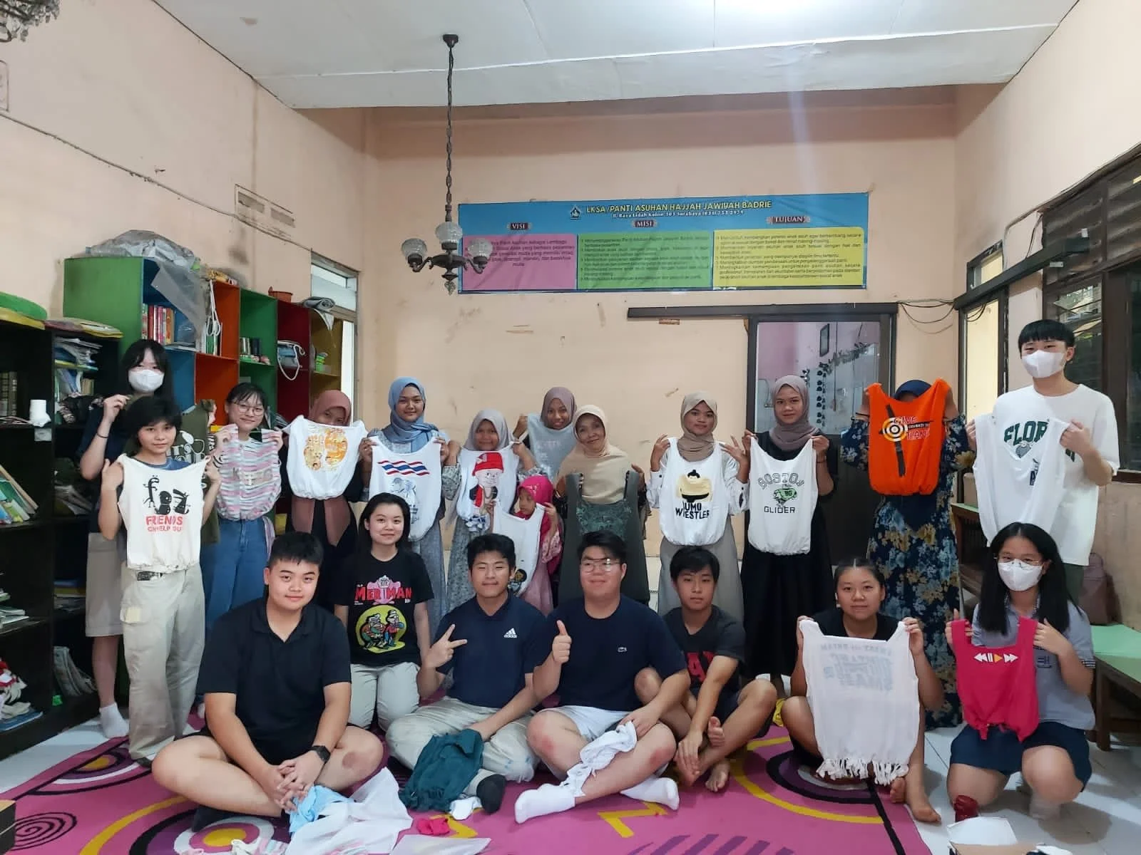 Group of children and adults standing indoors, some holding bags with designs, in a classroom with shelves, books, and a colorful poster on the wall.