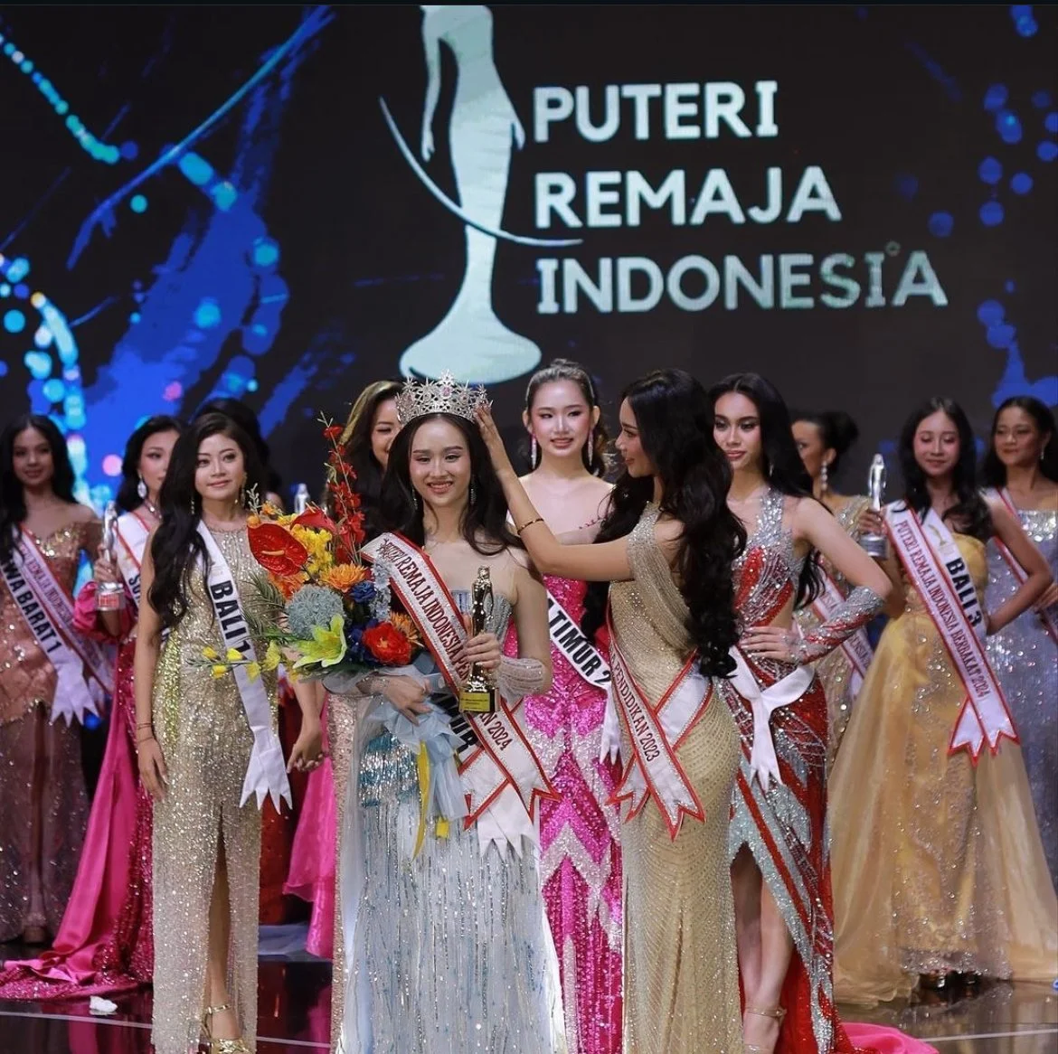 Group of women in formal gowns on stage, with a woman being crowned Miss Puteri Remaja Indonesia 2024, holding a bouquet and trophy, in front of a screen displaying a logo and text 'Puteri Remaja Indonesia'.