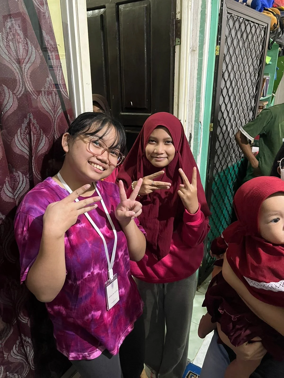 Two women smiling and making peace signs with their hands indoors. The woman on the left has glasses and is wearing a pink and purple tie-dye shirt with a lanyard. The woman on the right is wearing a maroon hijab and matching long sleeve shirt.