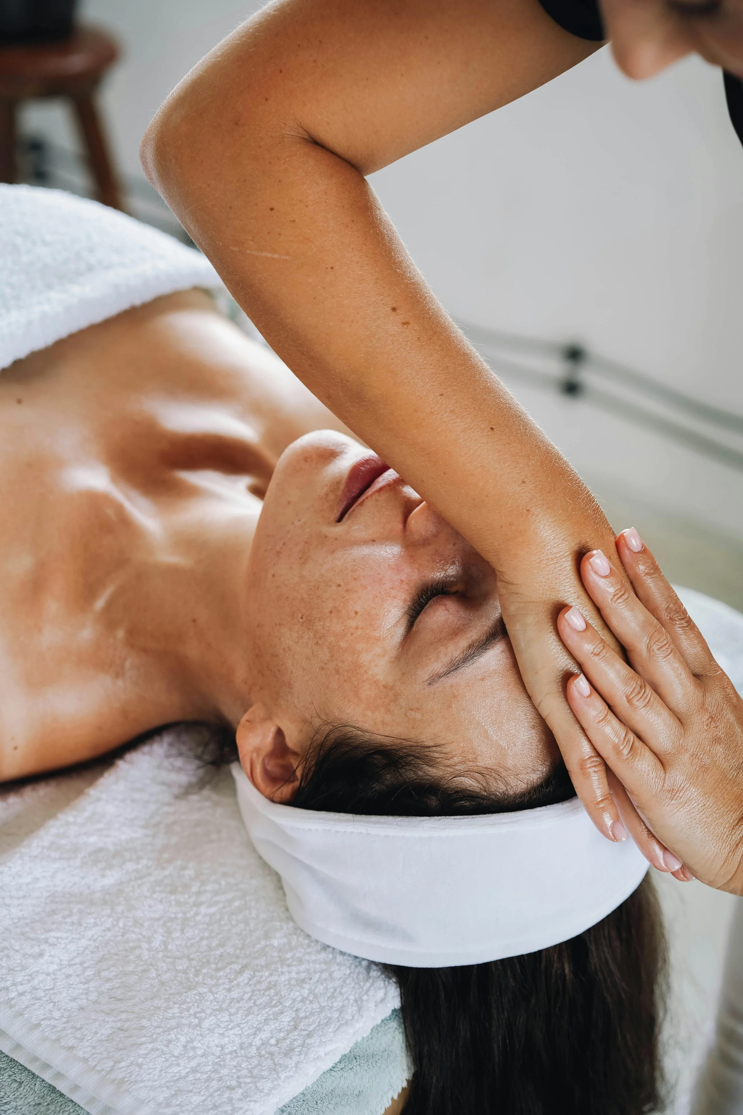 A woman receiving a facial massage at a spa, lying down with a white headband and towel on a massage table.
