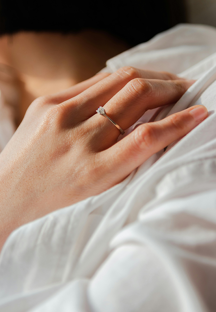 Close-up of a woman's hand with an engagement ring on her finger, resting on a white fabric surface.