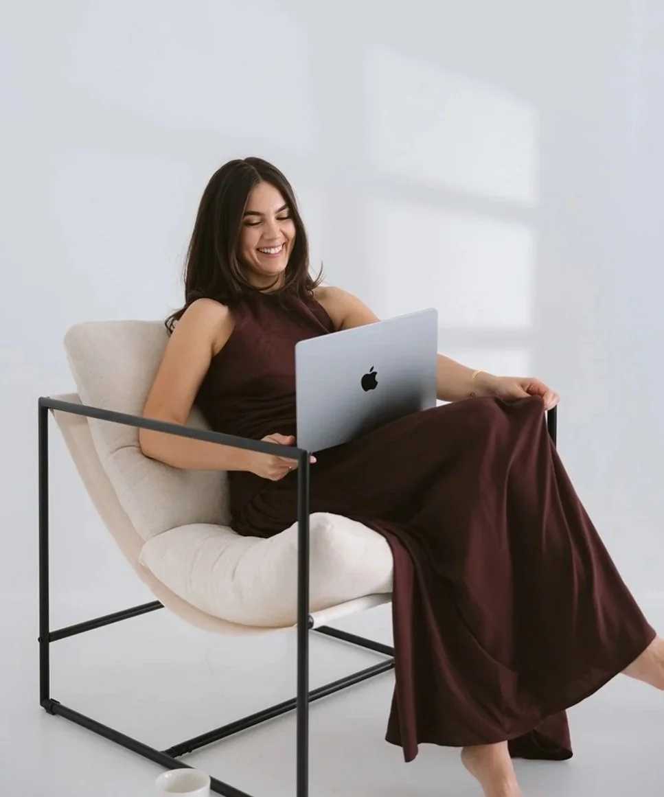 A young woman with dark hair and a sleeveless dress sitting on a modern chair with a cream cushion, smiling while using a silver Apple MacBook.