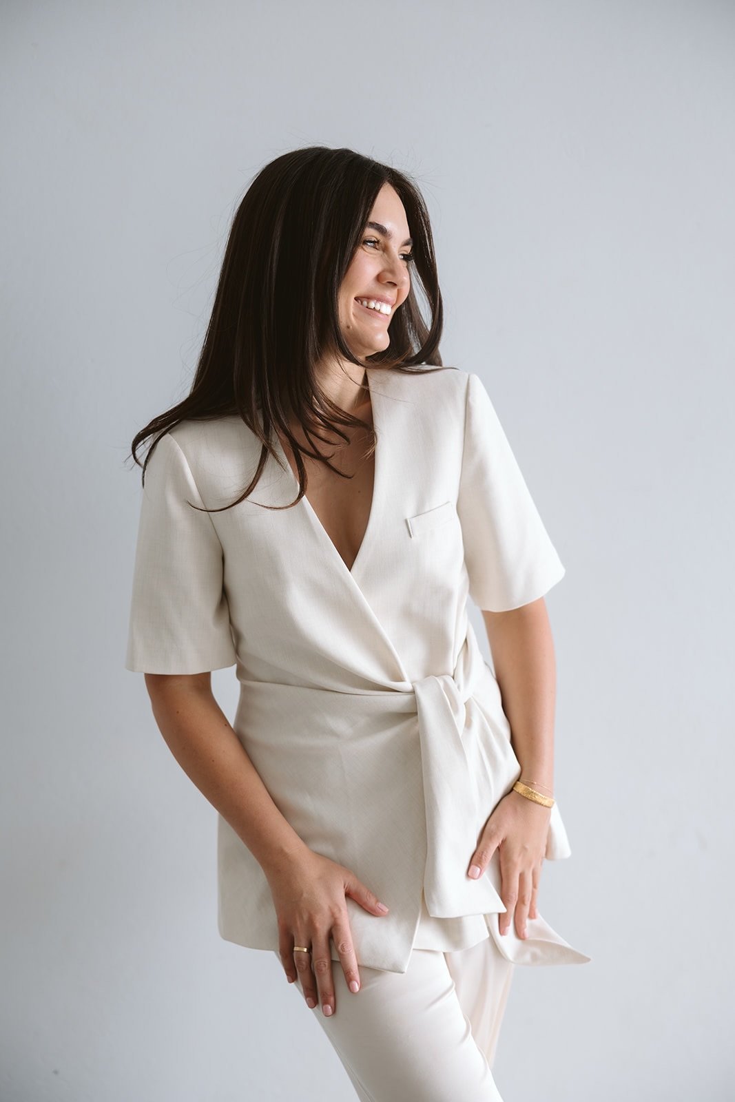 A woman with long dark hair wearing a cream-colored outfit, smiling and posing against a plain light gray background.