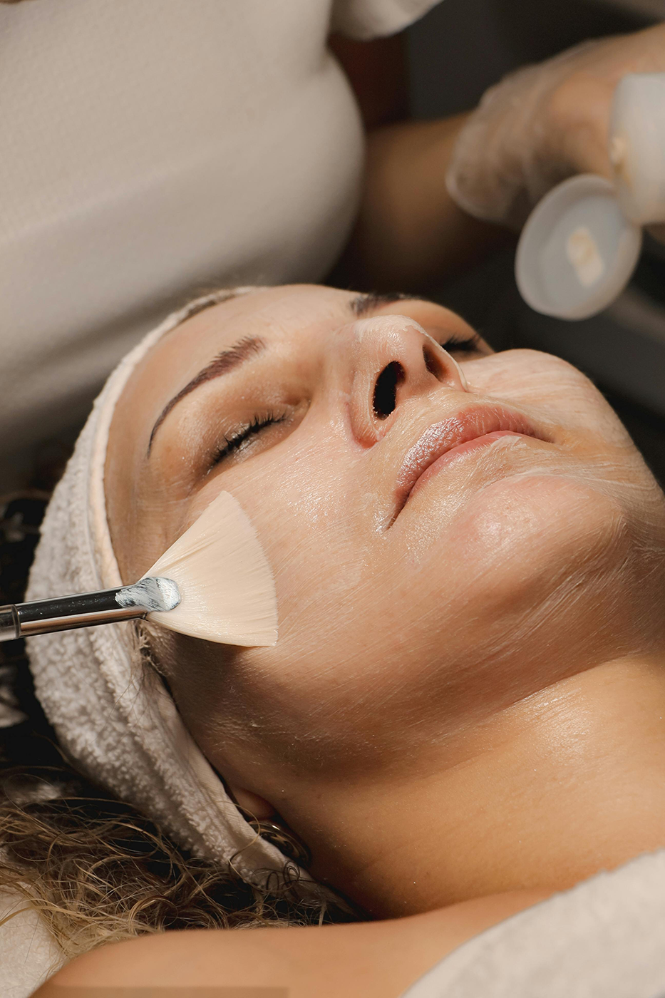 A woman lying down with eyes closed, receiving a facial treatment, with a brush applying skincare cream on her face.