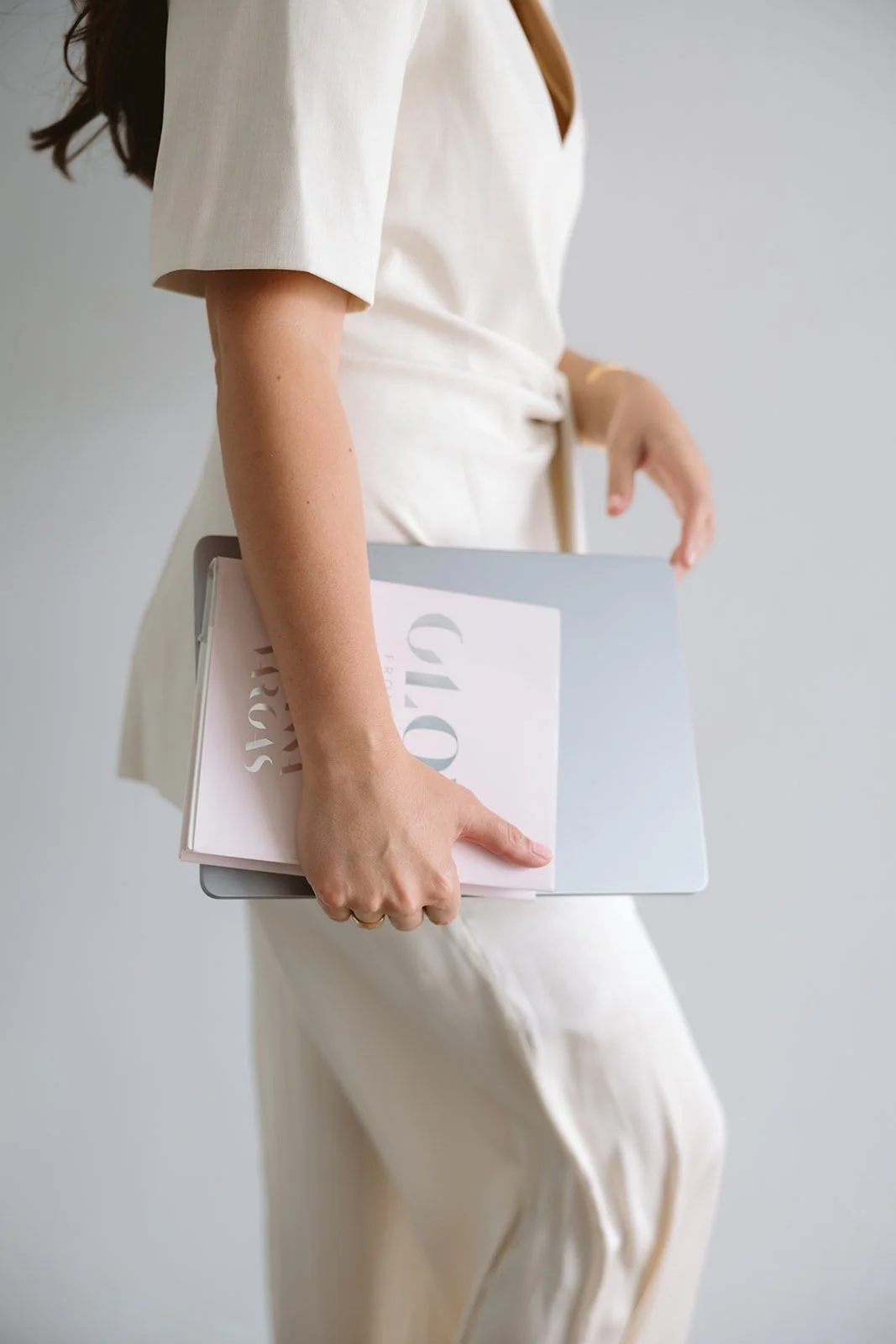 A woman in a cream-colored suit holding a laptop and a notebook.