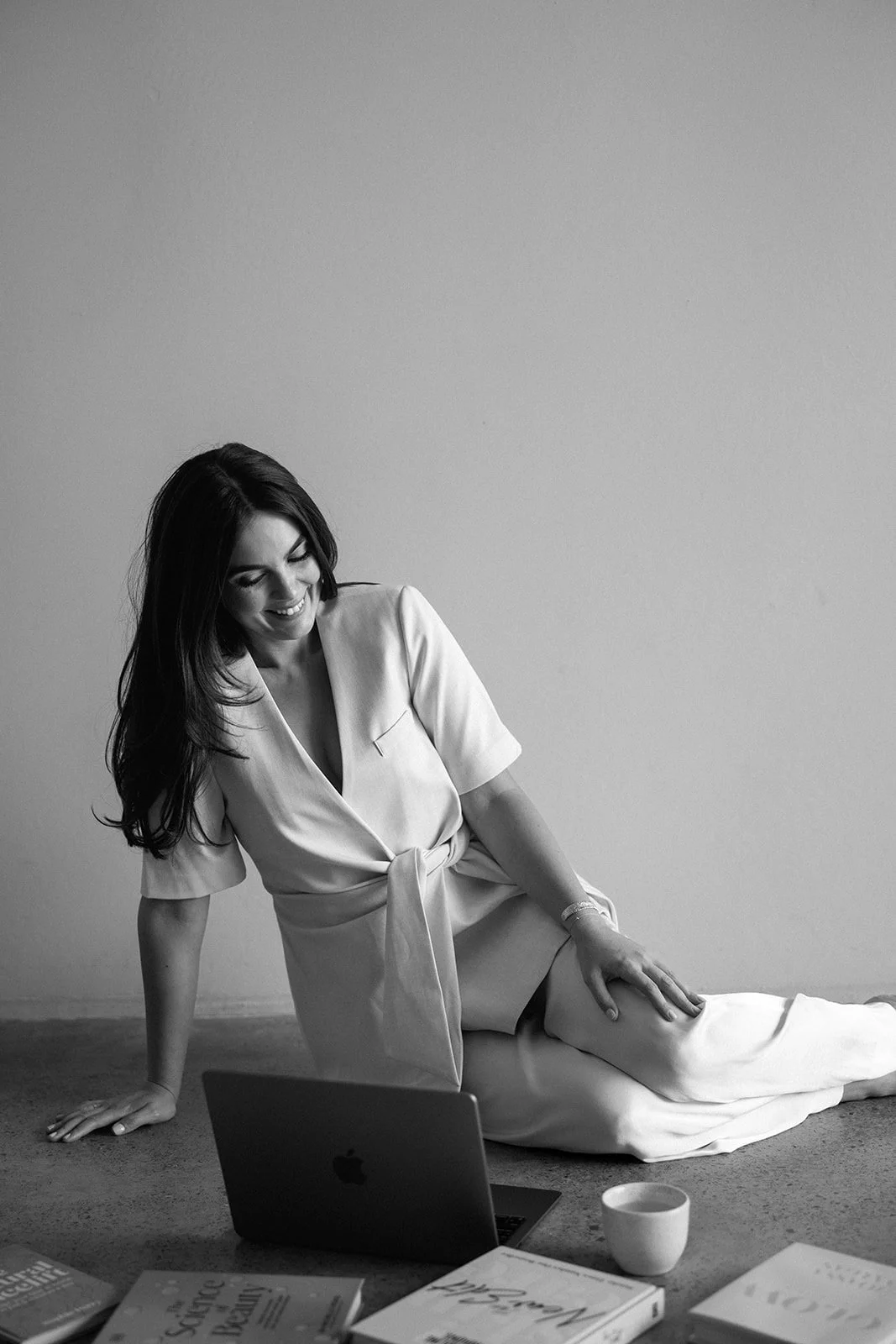 A woman sitting on the floor, smiling, with an open laptop, books, a coffee cup, and a magazine around her.
