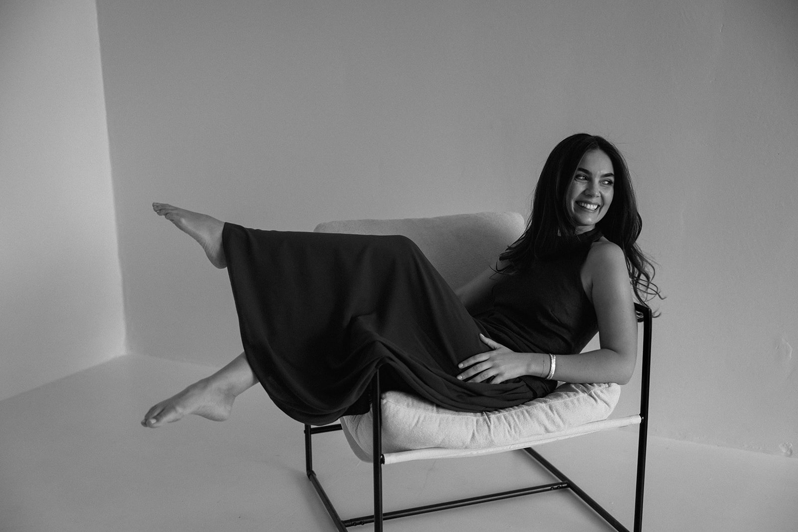 A woman in a black dress is lying on a modern chair with a cushion, turned on her side, smiling and laughing, with her right leg extended outward and her left arm resting on her hip. The wall behind her is plain and the image is in black and white.