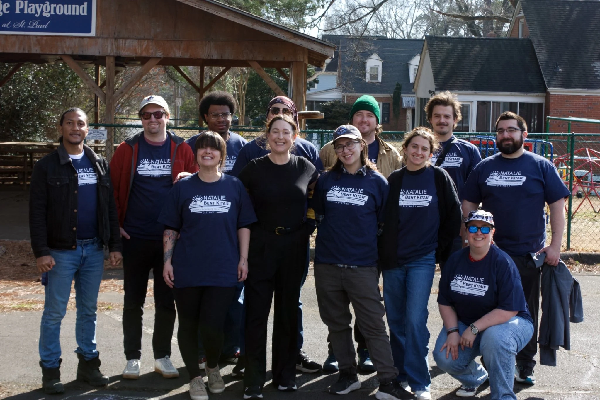 A group of volunteers wearing t-shirts that say "Natalie Bent Kitaif Durham Board of Education District 1" smile on a sunny afternoon after a successful canvas.