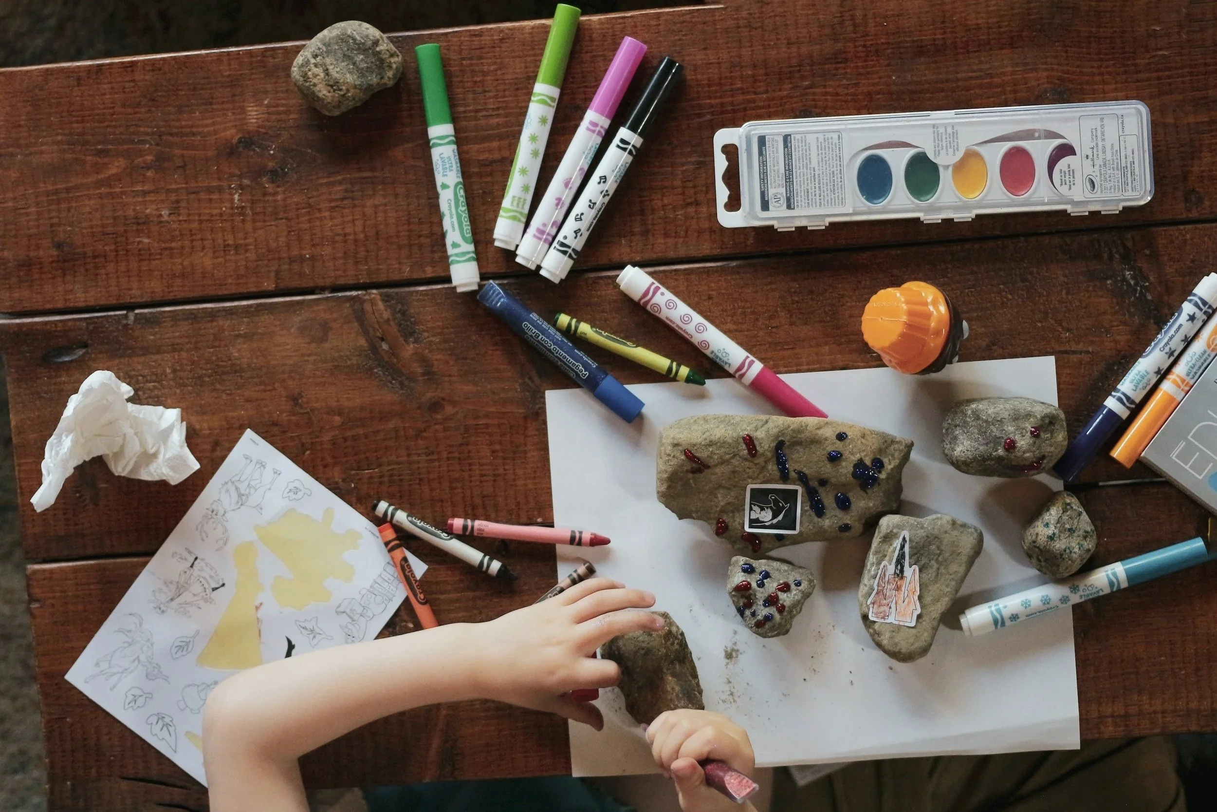 Child's hand carving a rock with a chisel surrounded by colorful markers, stones, and drawings on a wooden table.