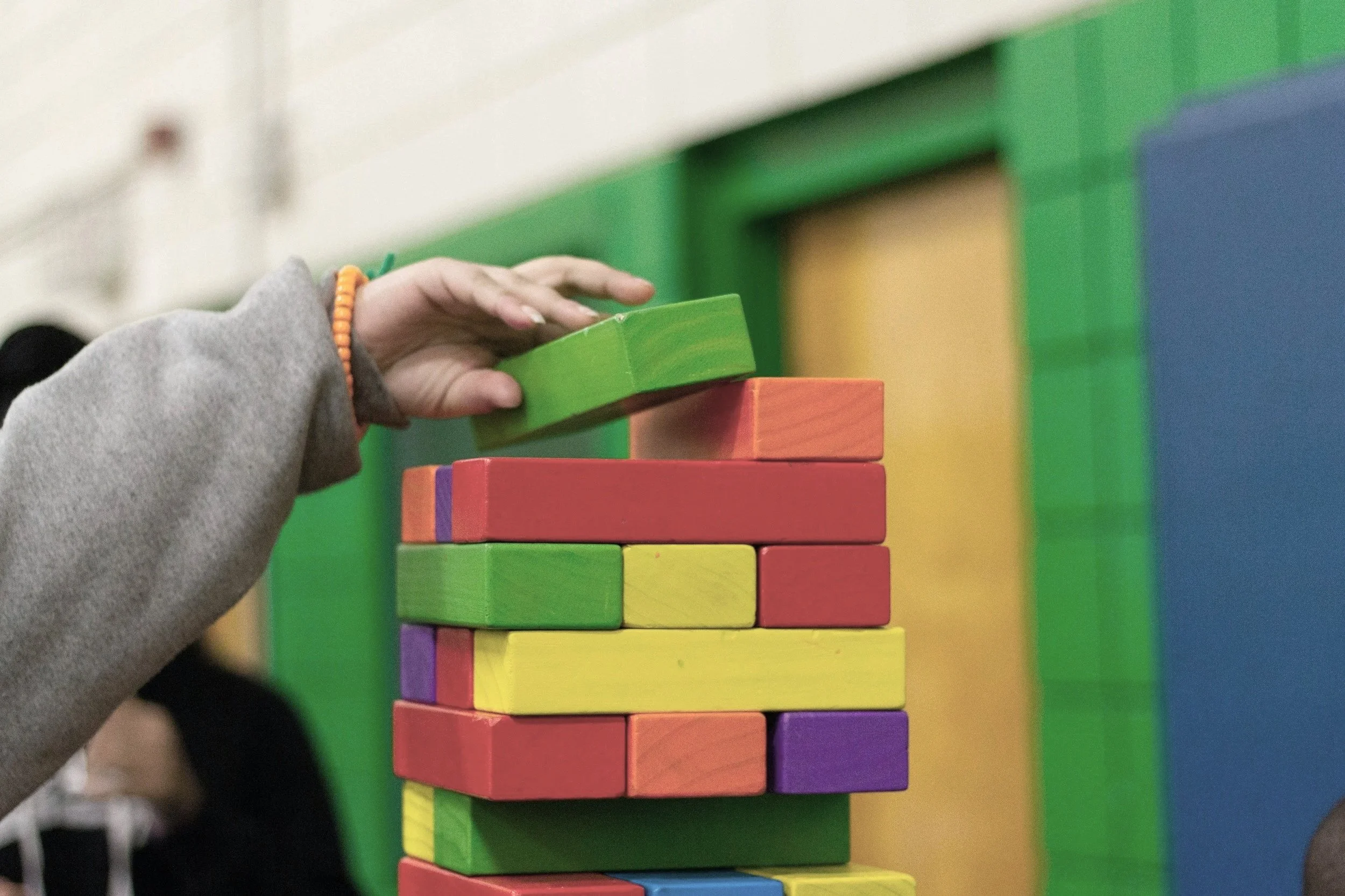 Close-up of a person's hand stacking colorful wooden blocks in a tower with a blurred background of green, yellow, blue, and orange sections.