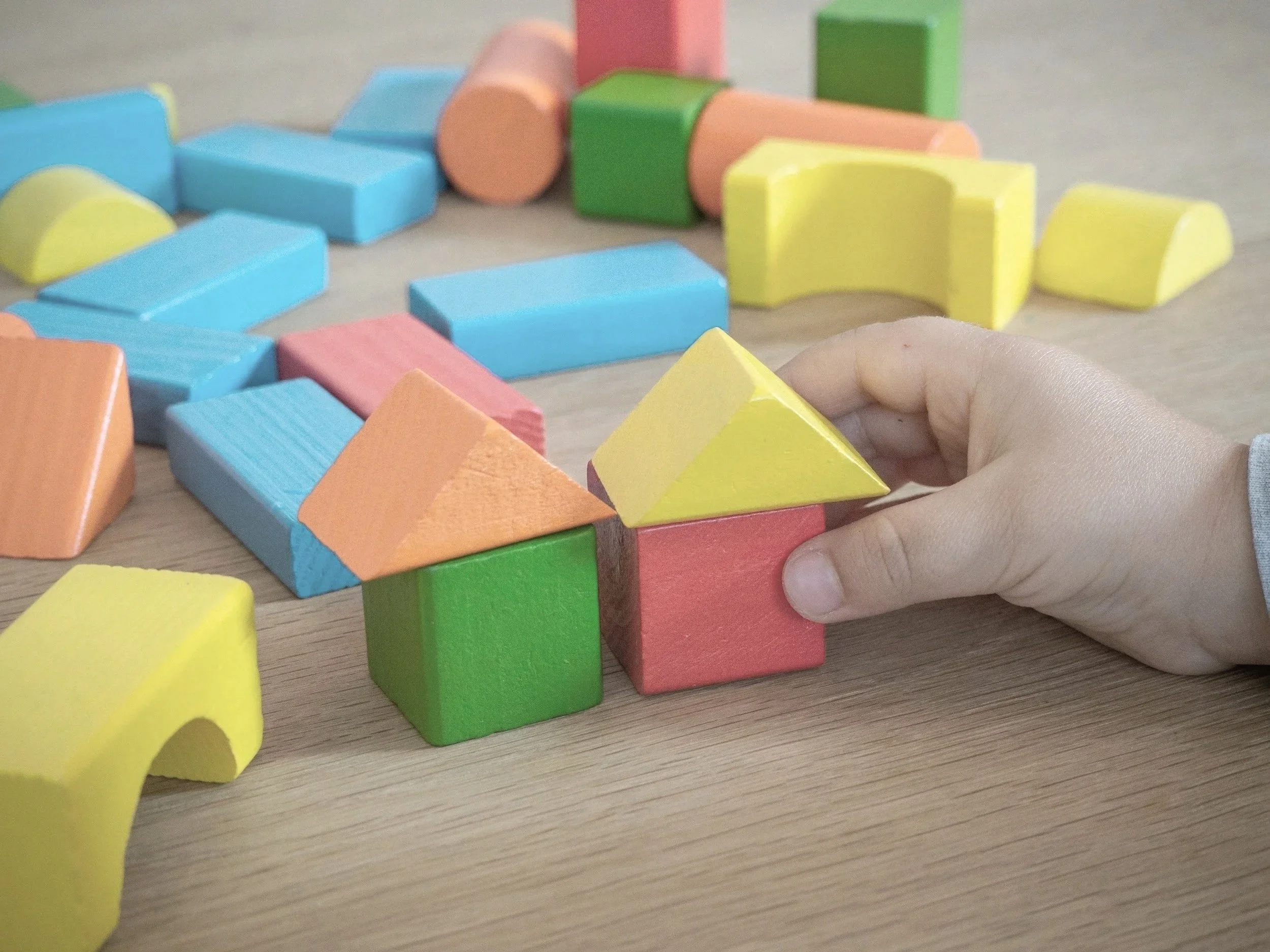 A child's hand arranging colorful wooden building blocks on a wooden surface, with assorted blocks scattered around.