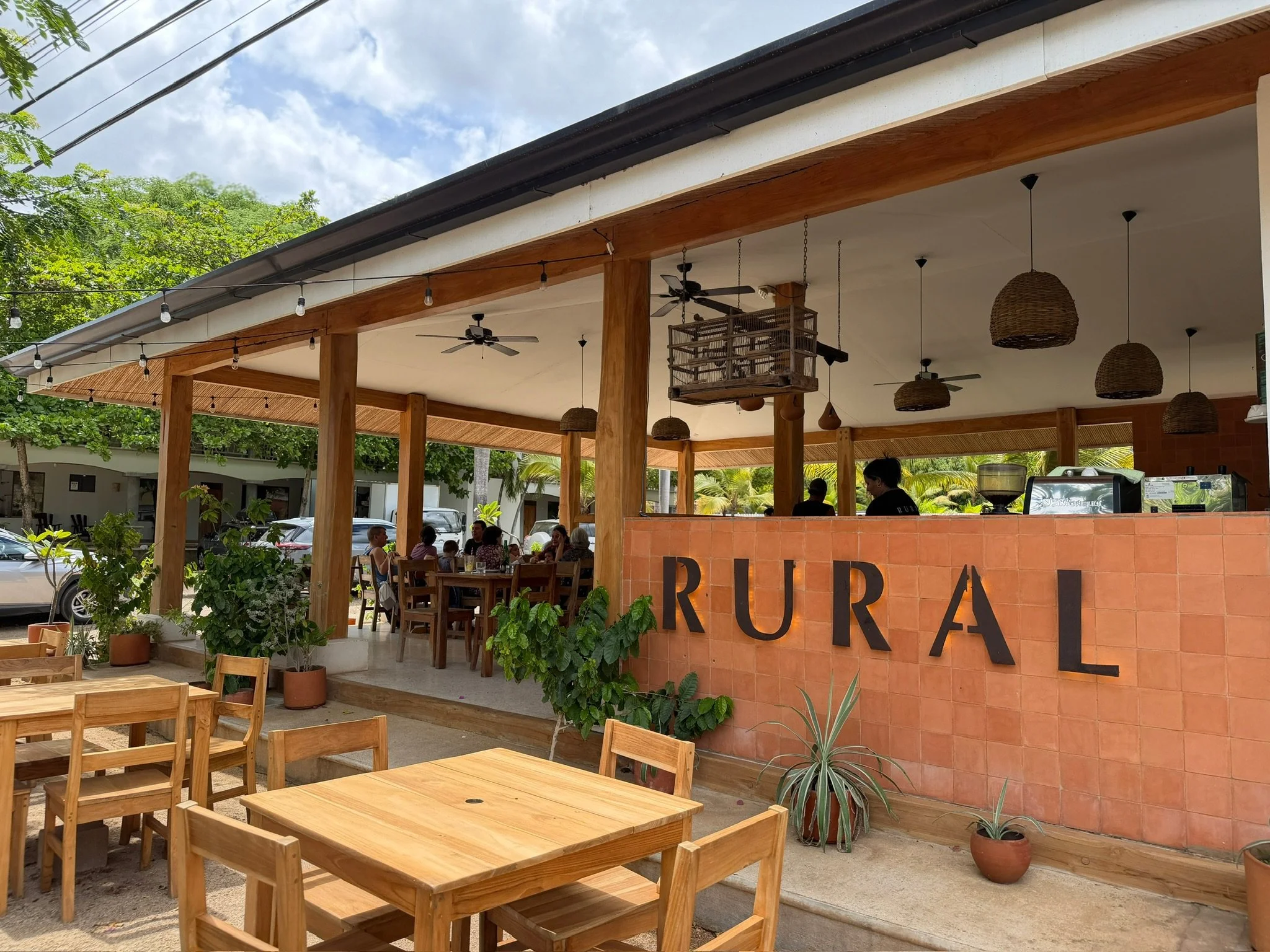 an open air restaurant with exposed wooden beams and string lights, simple wooden table and chairs outside and inside, and the letters RURAL on the front of the building