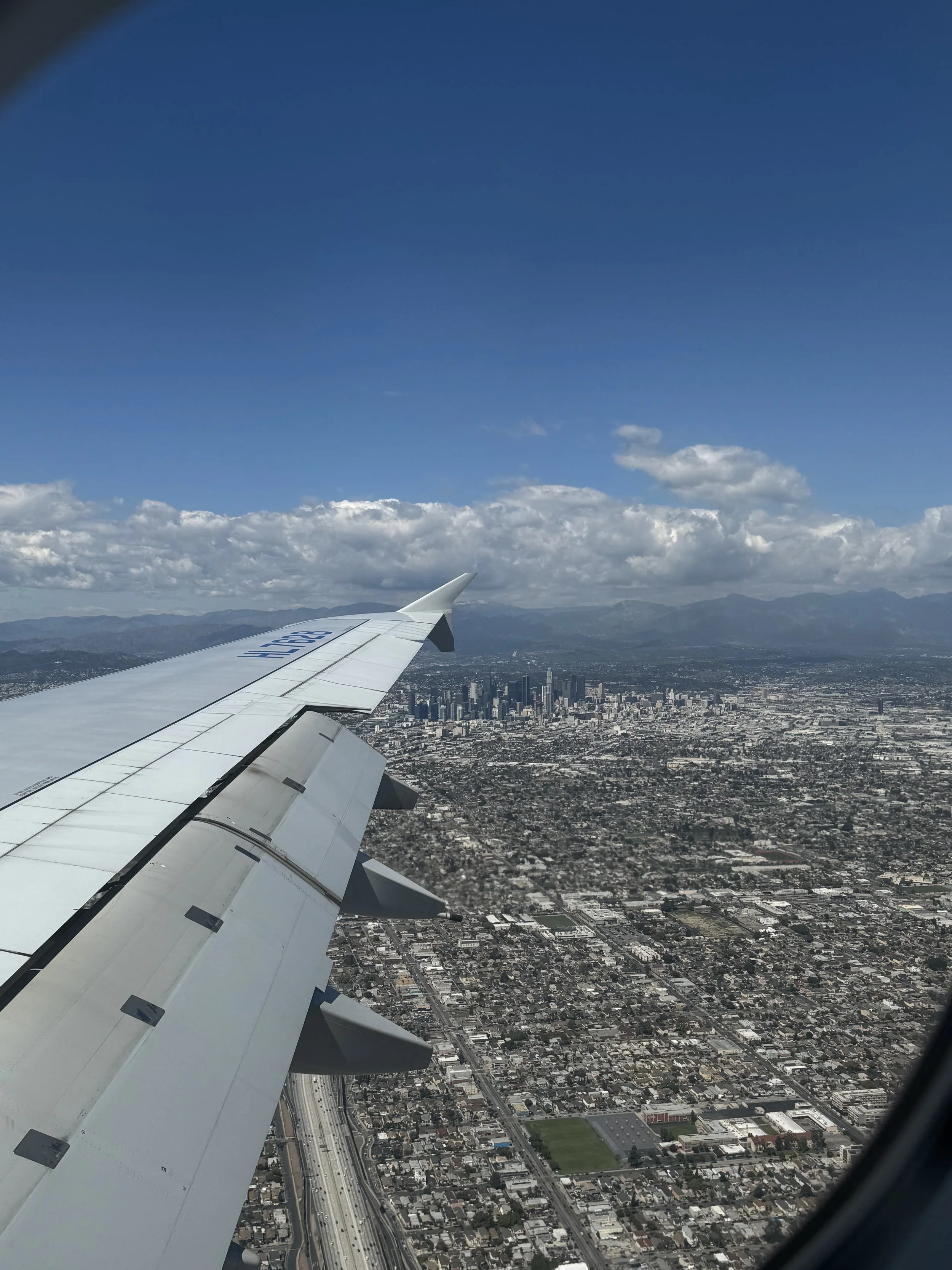 View of a city from an airplane window, showing the airplane wing and the cityscape below with buildings, roads, and surrounding mountains under a partly cloudy sky.