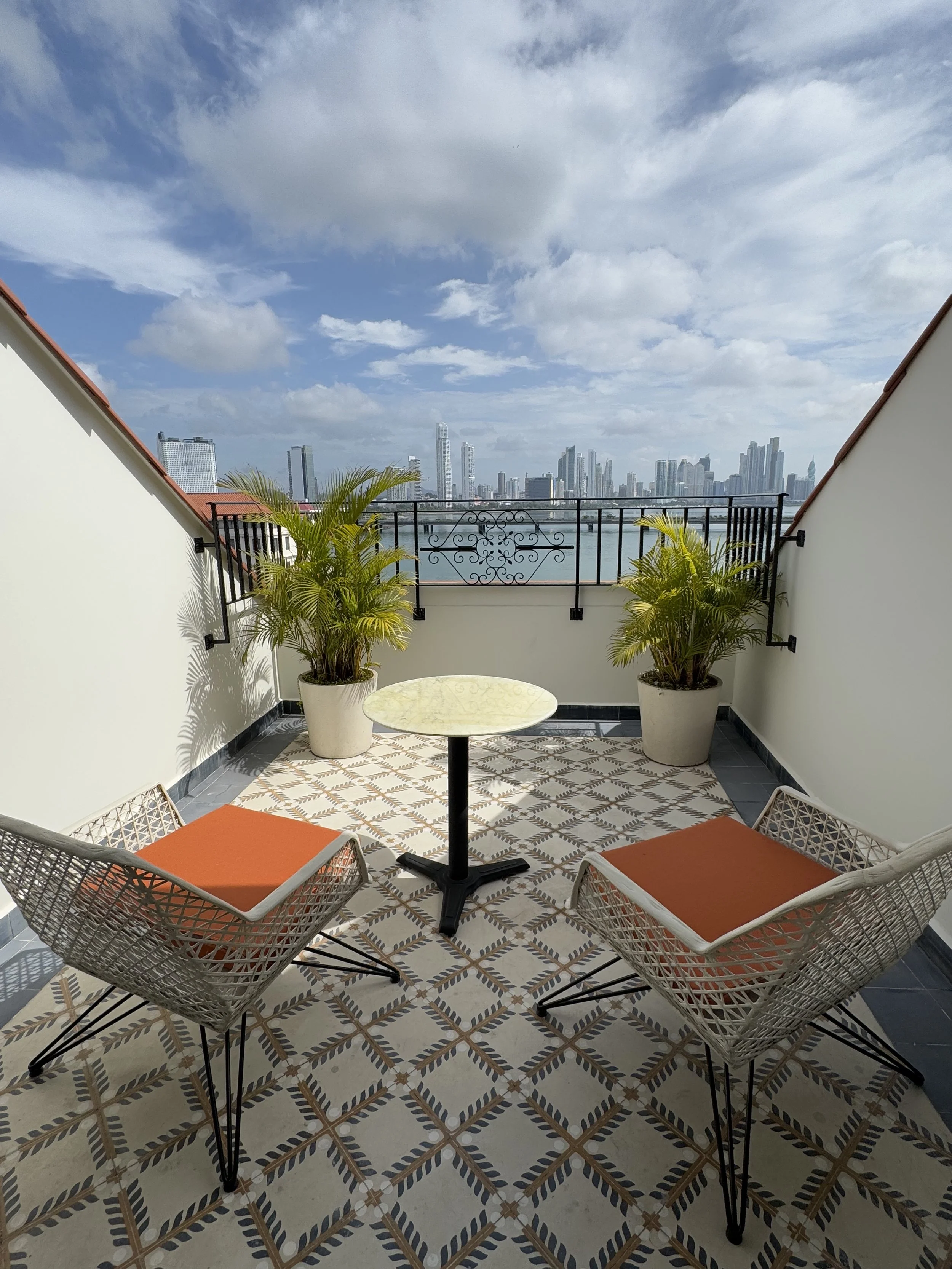Balcony along the water with two chairs with orange cushions, a round table, potted plants, and a city skyline in the background under a partly cloudy sky.