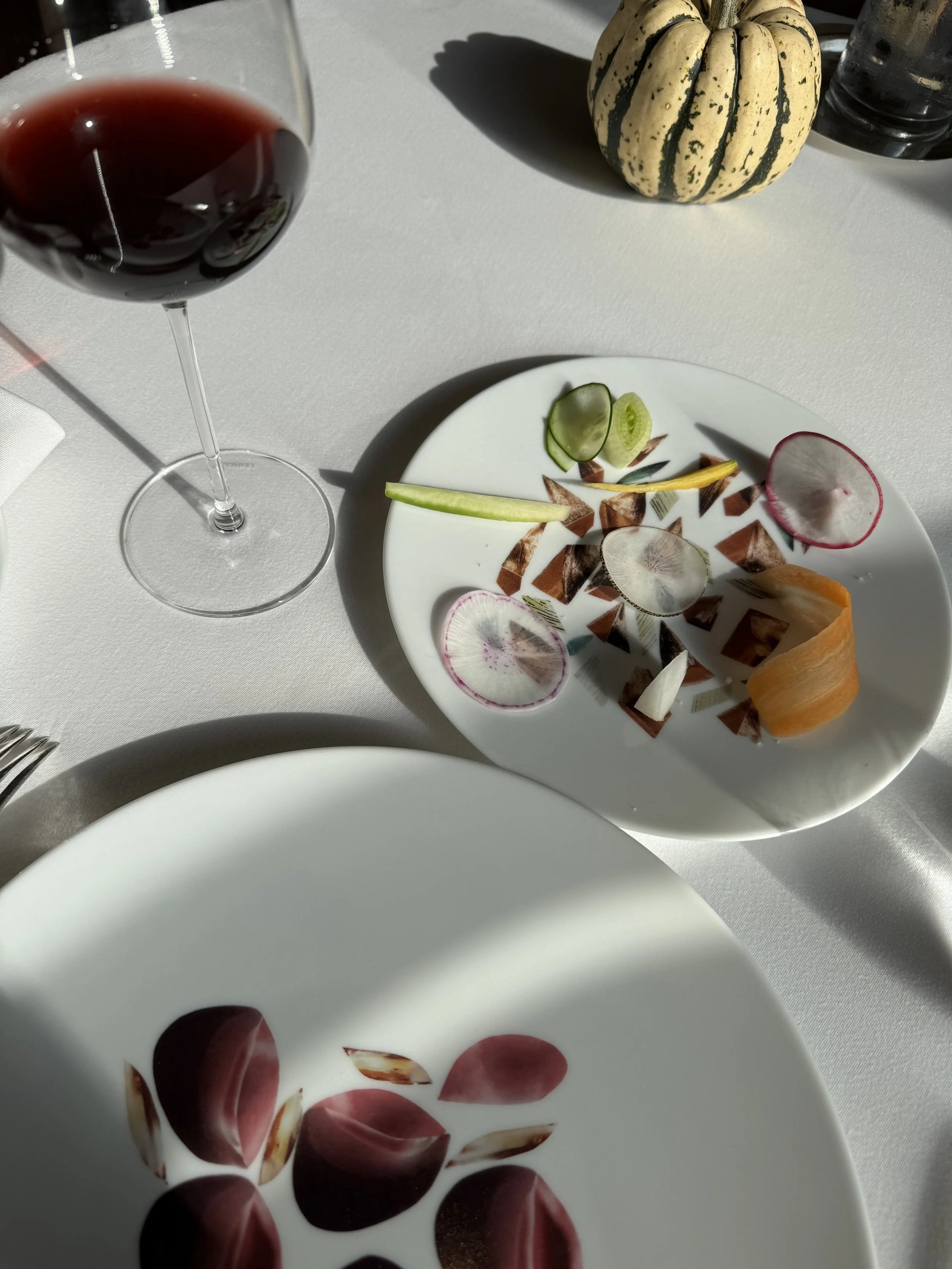 A plate of assorted sliced vegetables and radishes, a glass of red wine, a decorative pumpkin, and a glass of water on a white tablecloth.