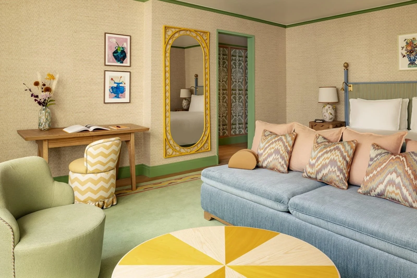 Colorful hotel room with a green armchair, a blue sofa with patterned pillows, a round yellow and white table, a wooden desk with a chevron patterned chair, wall art, a vase with flowers, and a mirror reflecting a bed.