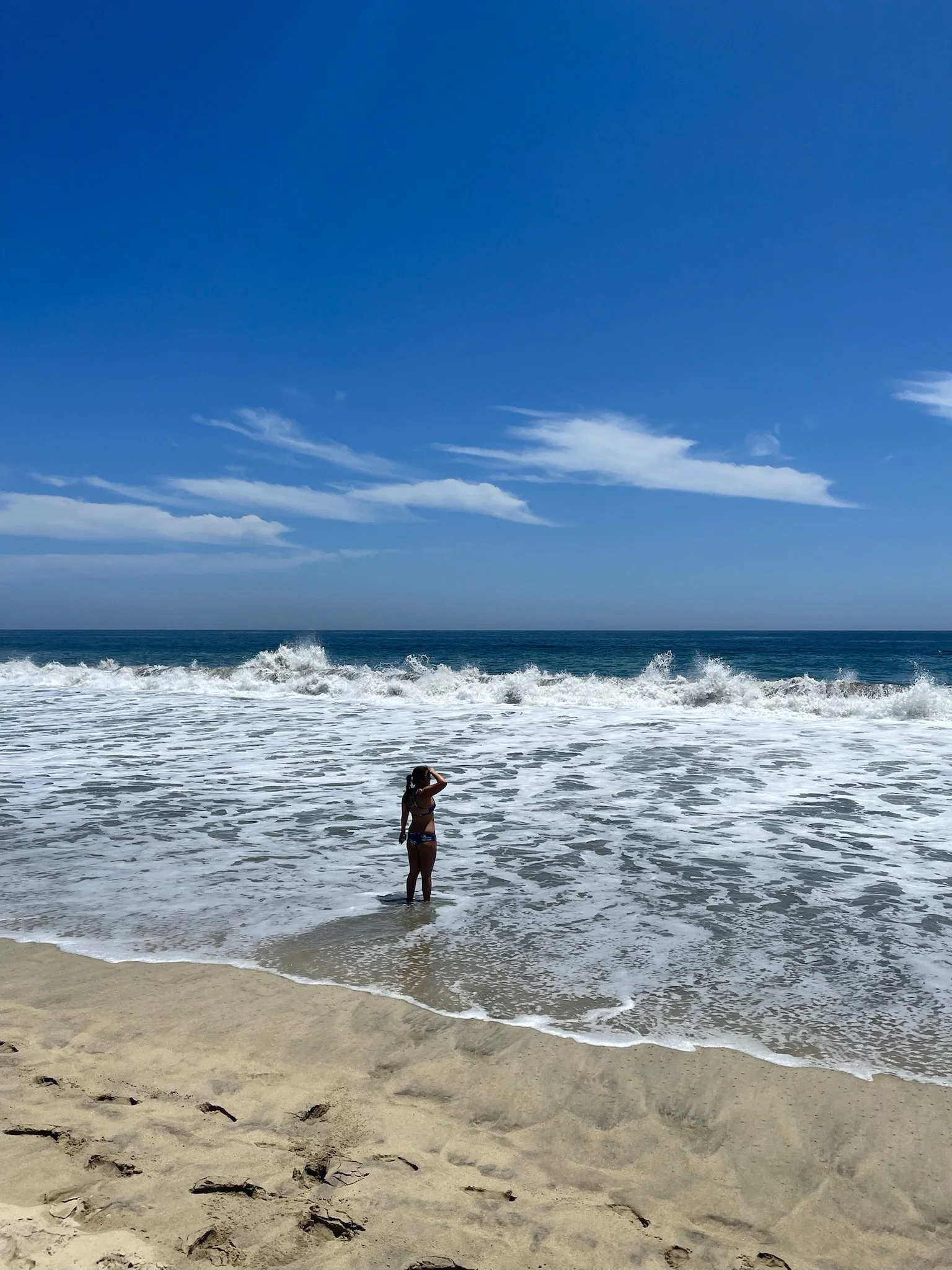 A woman standing in the shallow water of a sandy beach, looking at the ocean under a blue sky with a few clouds.