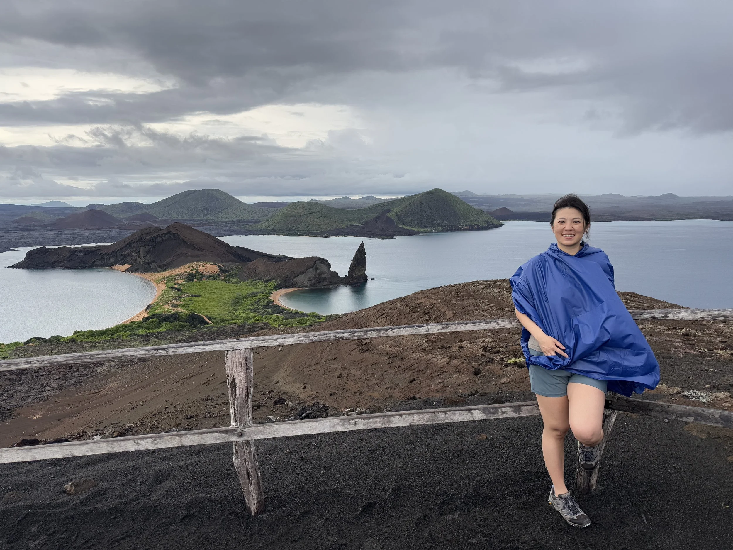 A woman in a blue rain poncho and shorts is standing on a viewing platform overlooking a volcanic crater lake with dark volcanic rocks and green vegetation, under a cloudy sky.