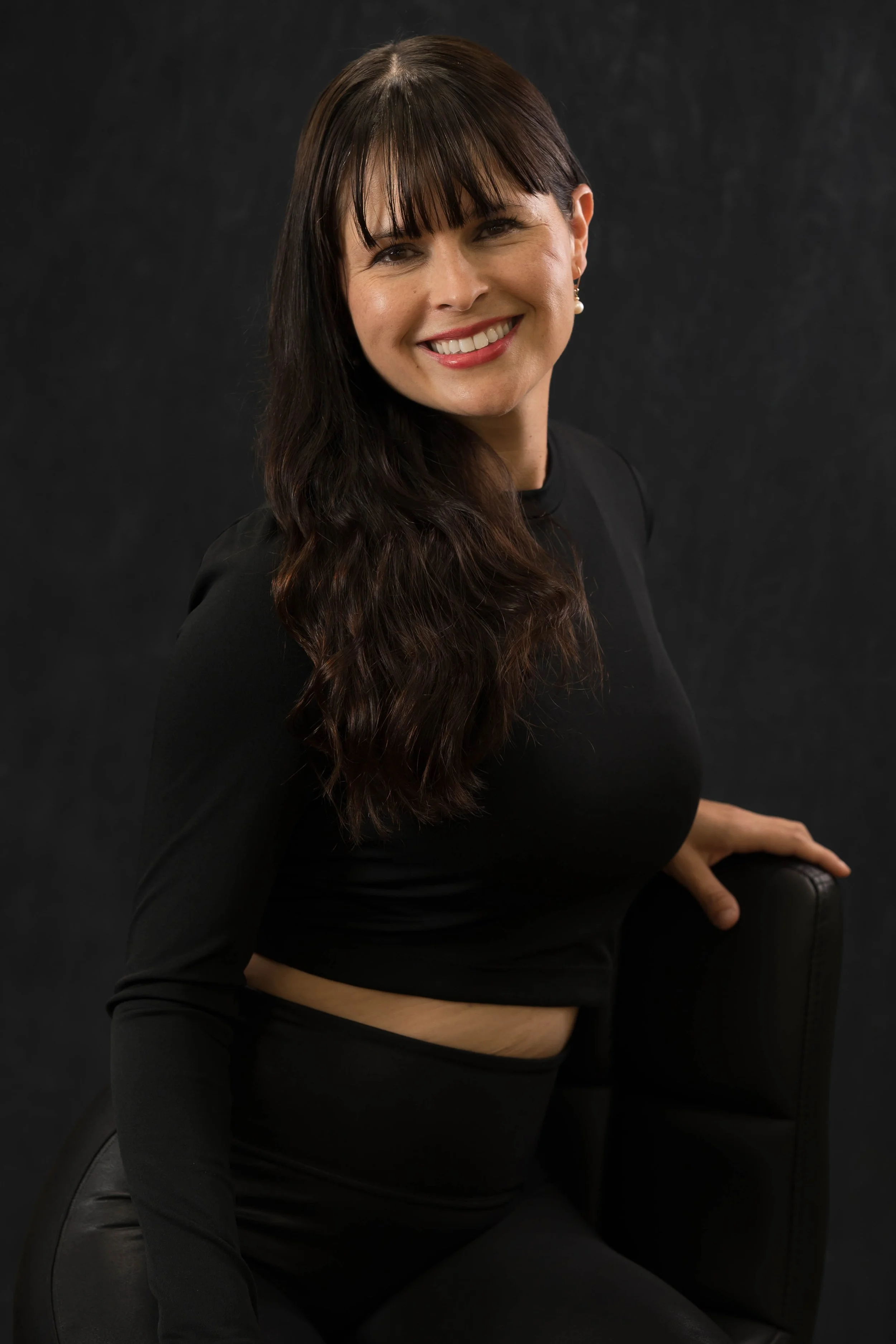 A woman with long, wavy, dark brown hair and bangs, smiling, dressed in black, sitting in front of a dark background.