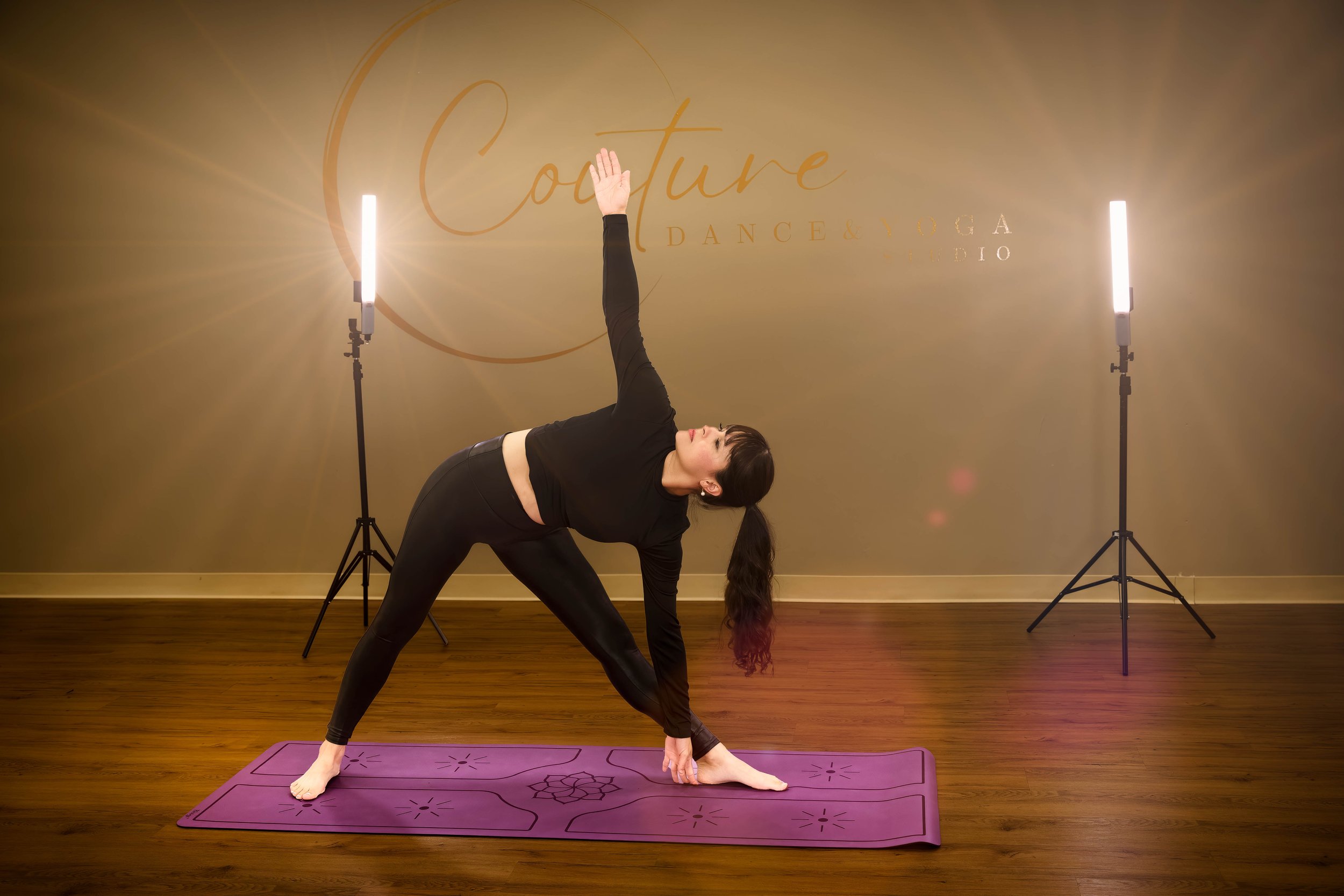 Woman practicing yoga on purple mat in studio with beige walls, two vertical lights, and 'Culture Dance & Yoga Studio' sign, wearing black activewear, in a lunge with one arm reaching up and the other hand on the floor.