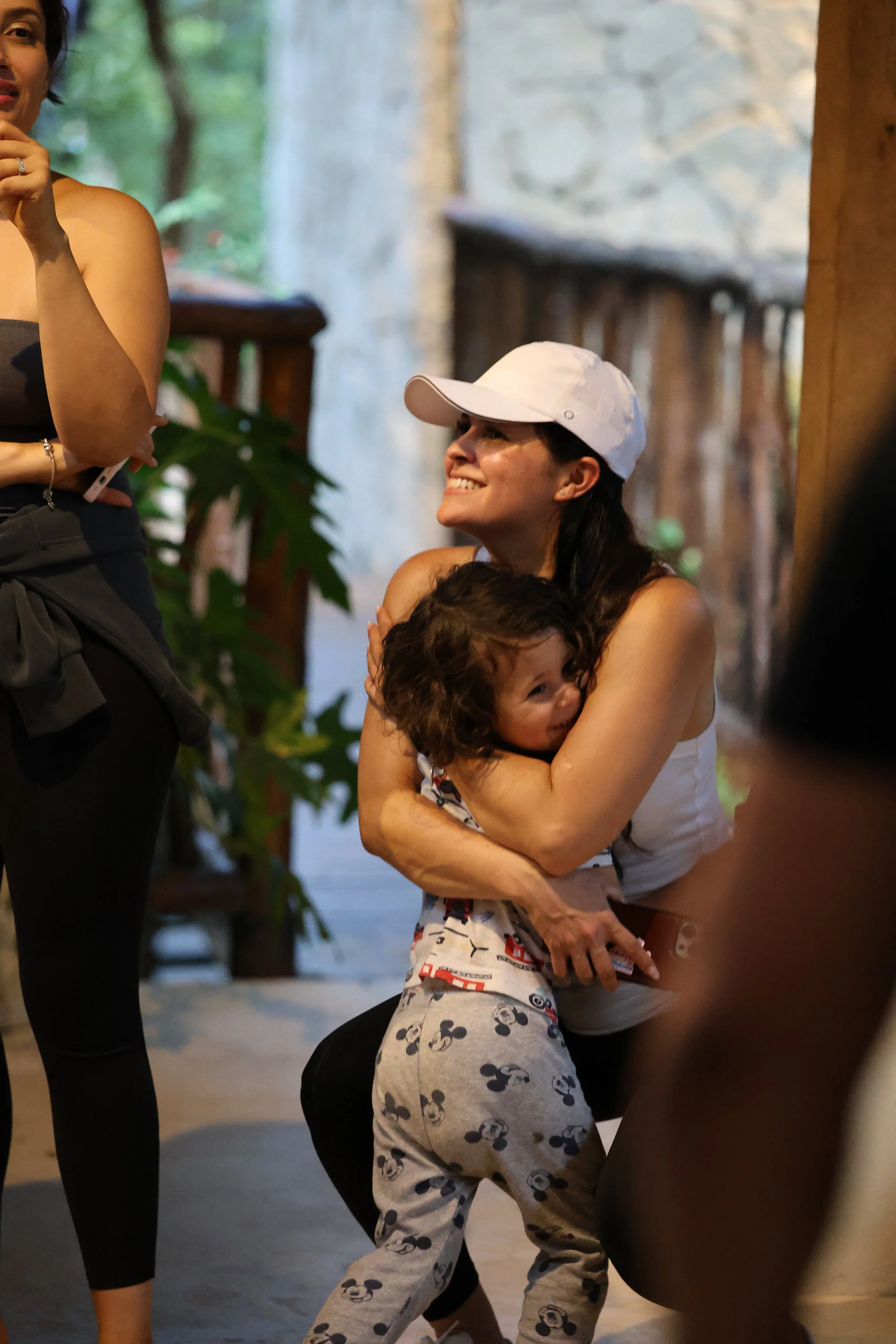 A woman wearing a white baseball cap is smiling and hugging a young boy who is wearing Mickey Mouse pajamas, inside a rustic setting with wood and stone decor.